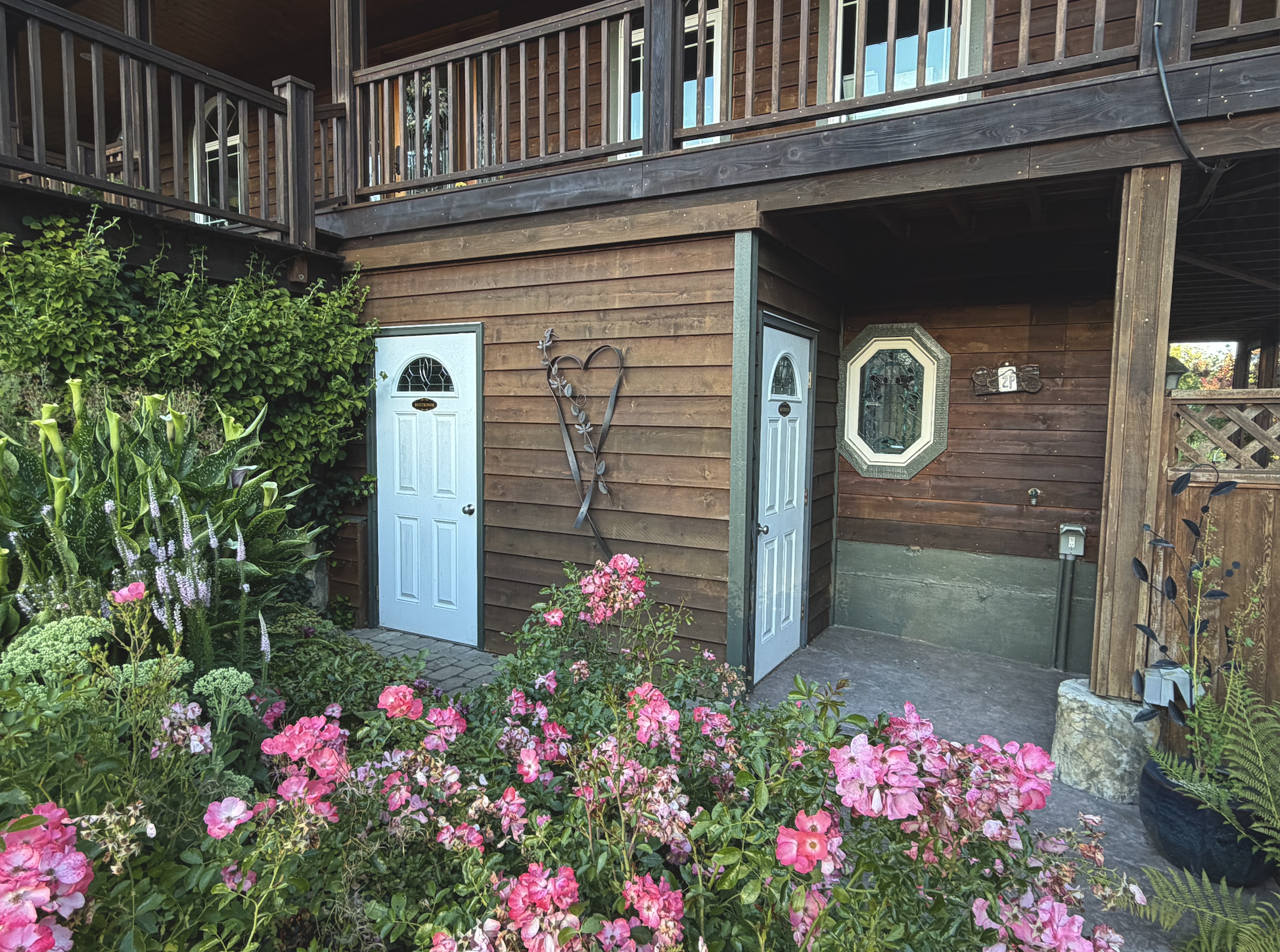 A two-story wooden house with a balcony, lush garden with pink flowers, and exterior doors