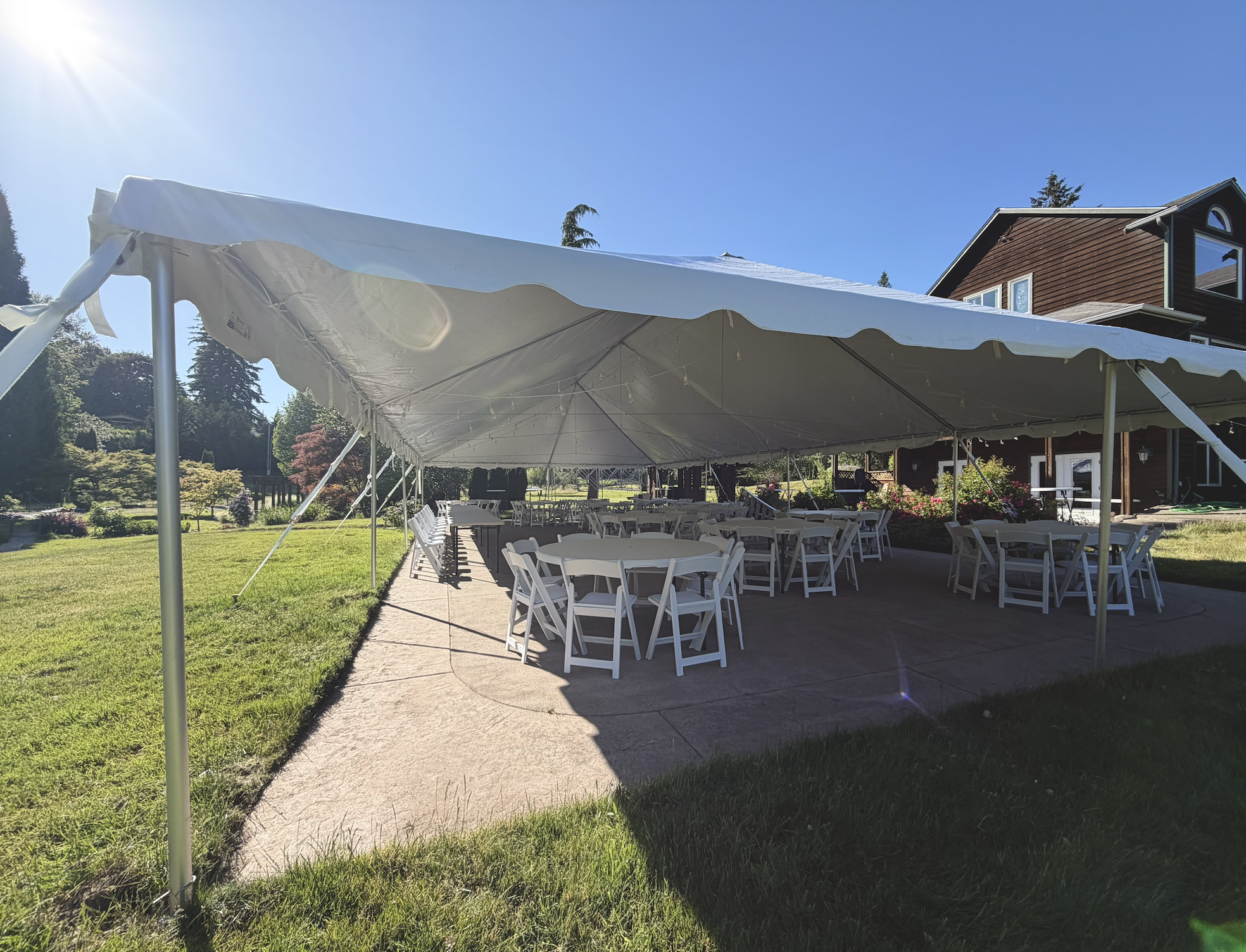 Outdoor setup with a large white canopy tent, round tables, and chairs on a grassy area adjacent to a house.