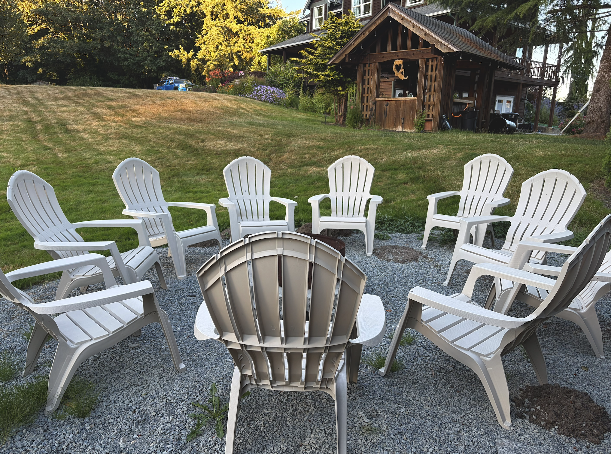 A circle of white plastic outdoor chairs arranged on a gravel surface in a backyard, with a wooden shed, grassy hill, trees, and a house in the background.