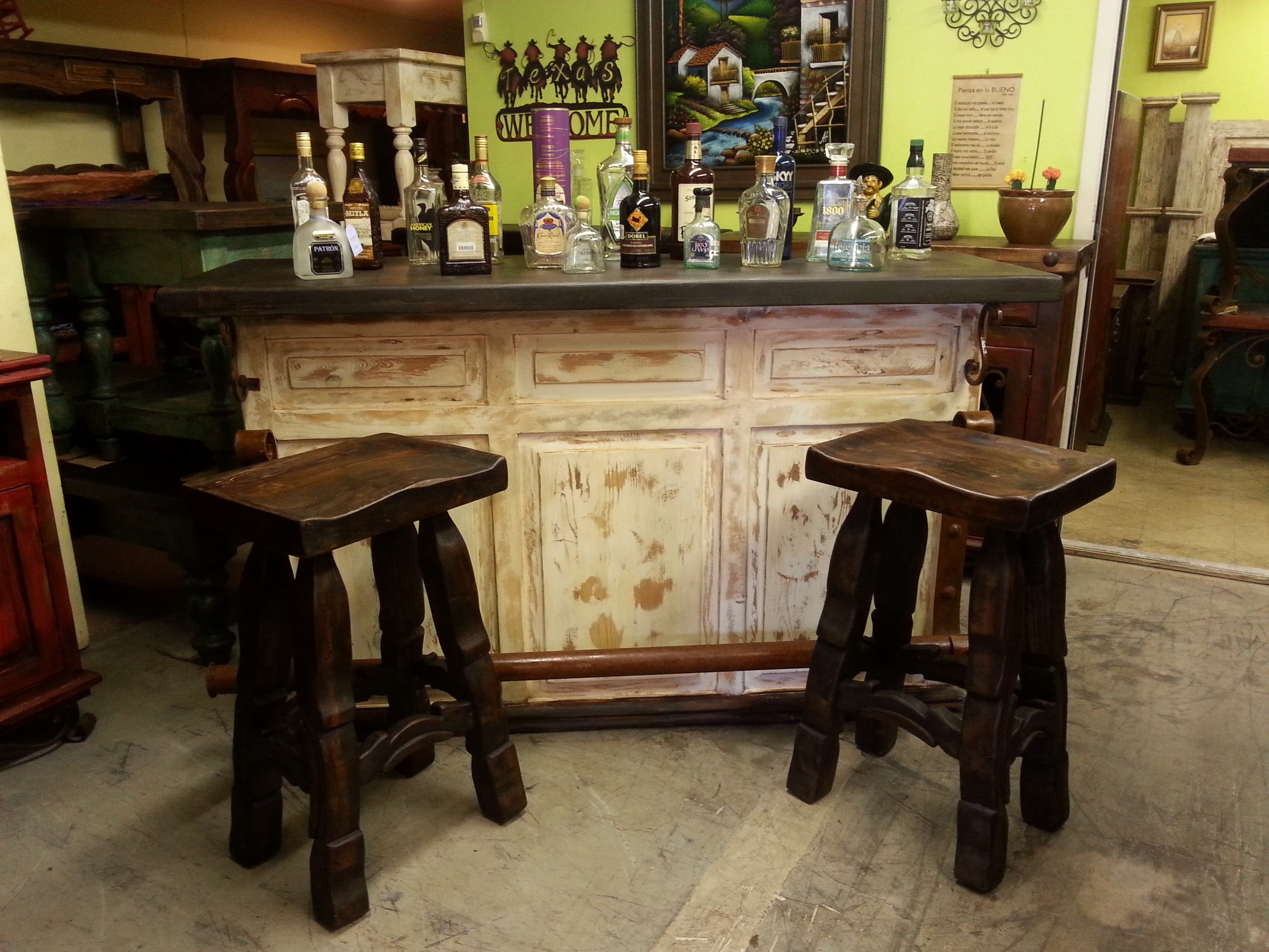 A rustic wooden bar with multiple bottles of liquor on top, set in a cozy, wood-themed interior with wall art, a welcome sign, and decorative plants.