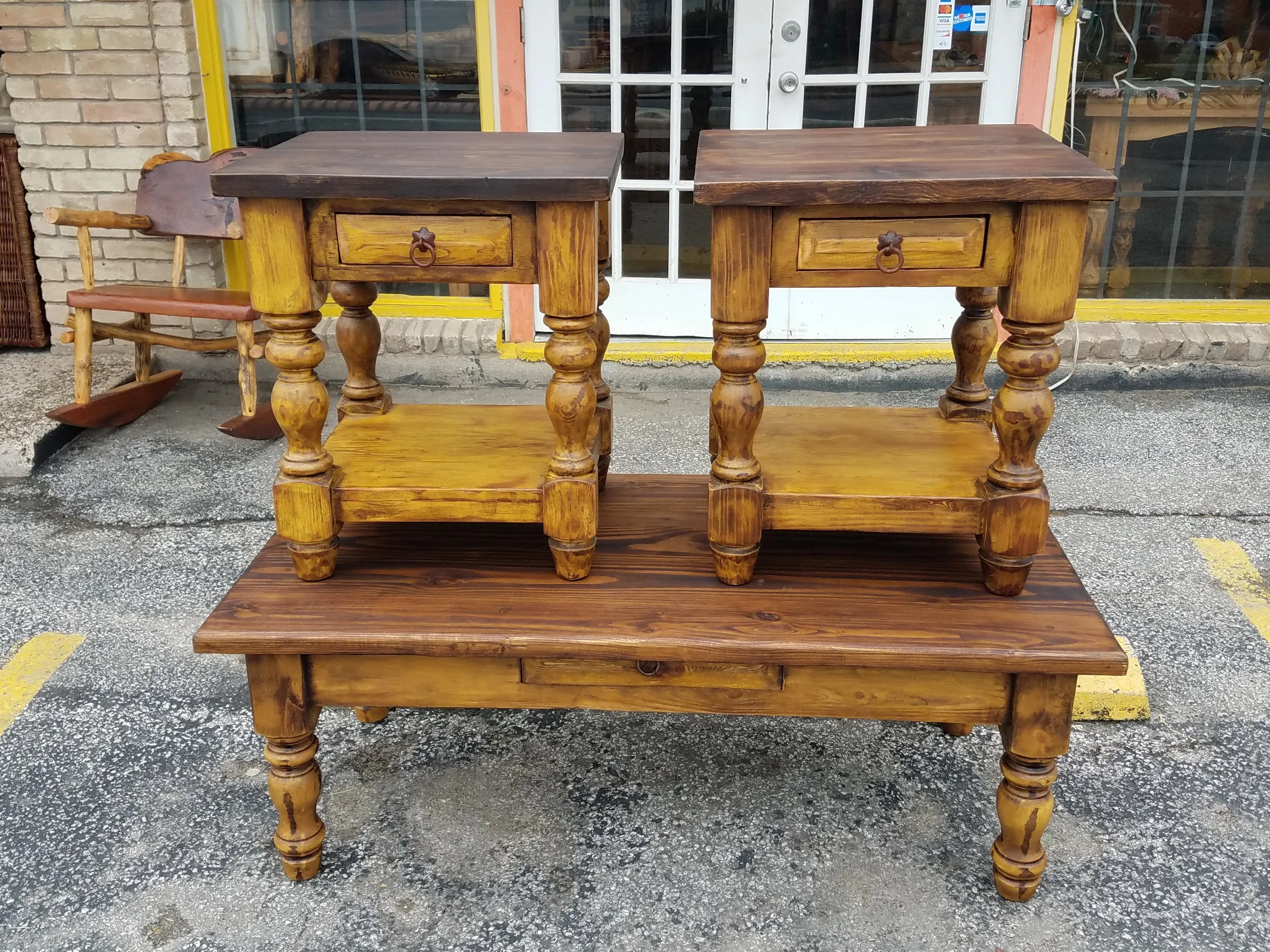 Two vintage wooden tables with drawers and turned legs placed outdoors on a concrete sidewalk in front of a storefront with glass doors and windows. The storefront has yellow trim around the door frame.