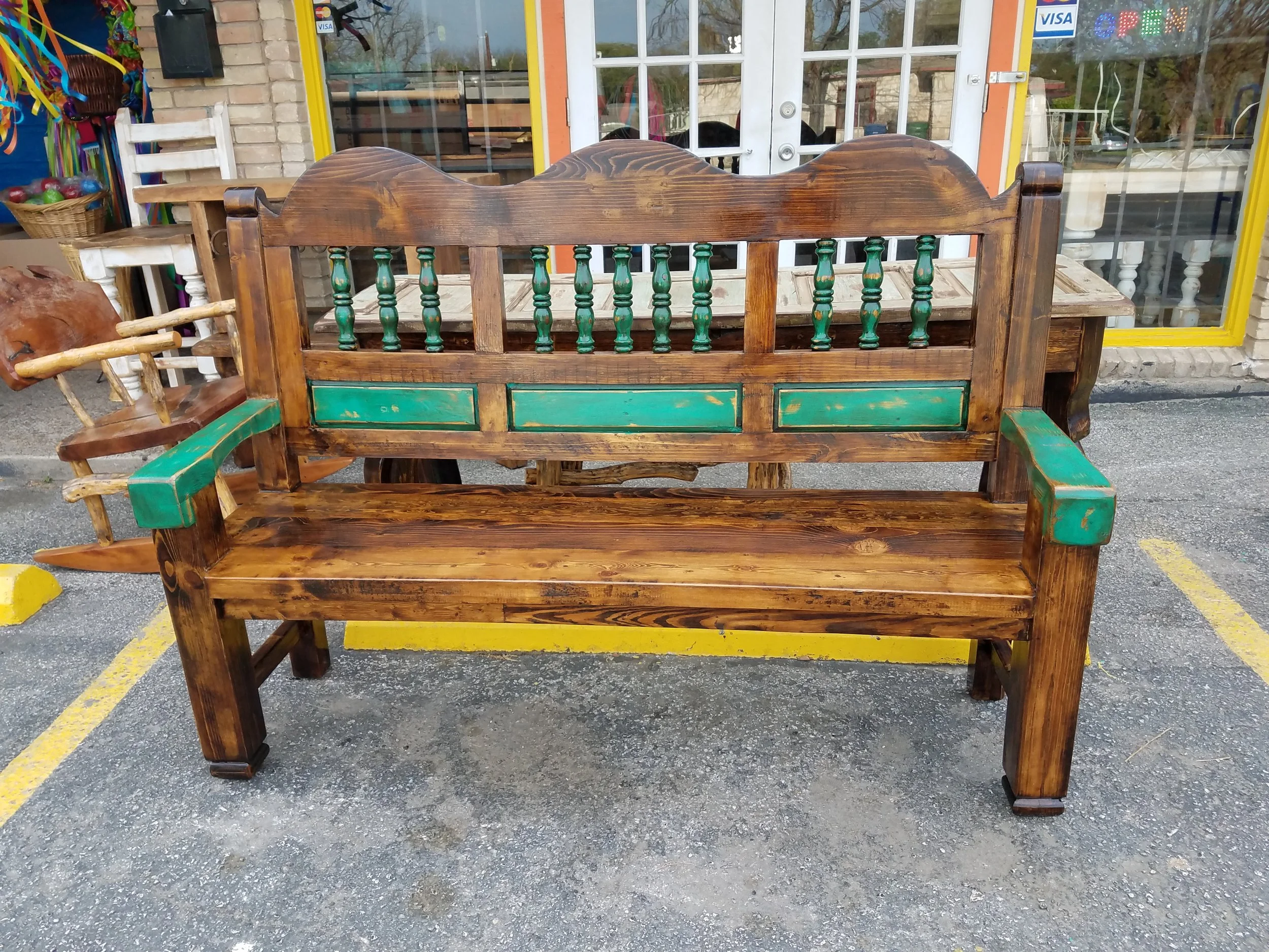 A wooden bench with green painted details is placed outside a storefront with a glass door. Various items and decorations are visible in the background.