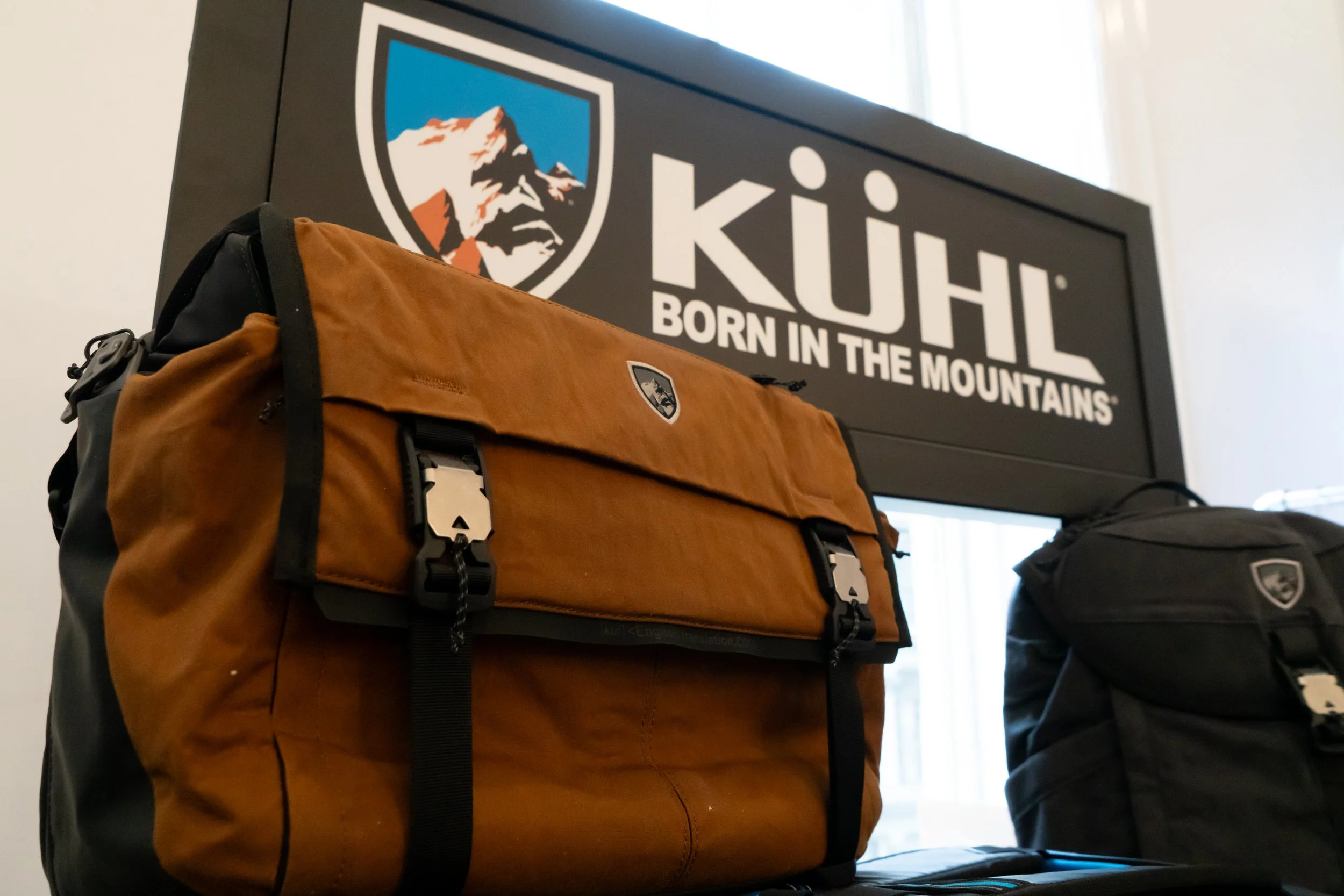 Kuhl backpacks displayed in front of a sign with the Kuhl logo and slogan 'Born in the Mountains' in an indoor setting.
