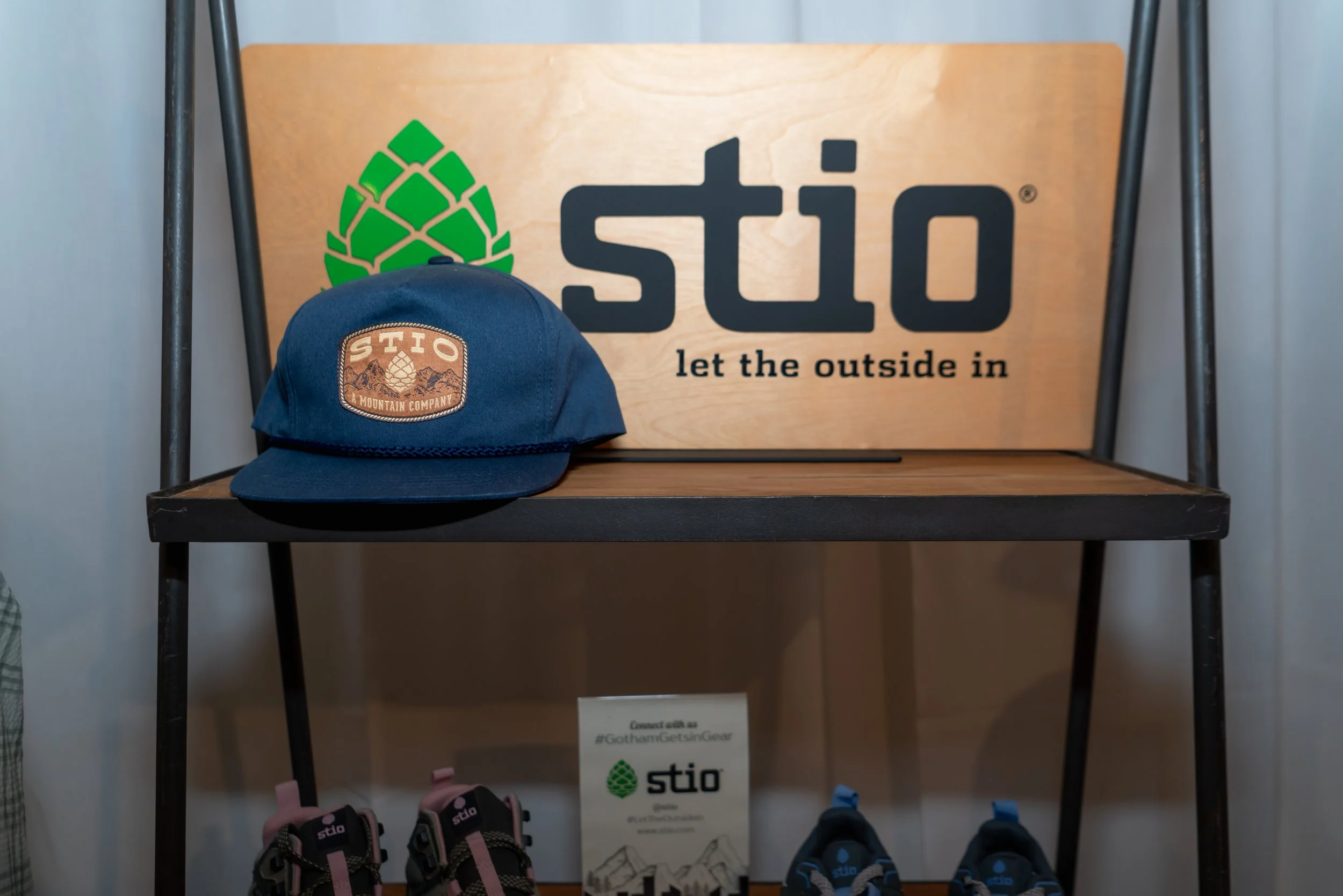 A navy blue cap with a brown patch that has the 'STIO' logo, placed on a wooden and metal display table. Behind it is a wooden sign with the 'stio' logo, the pine cone symbol, and the phrase 'let the outside in.'