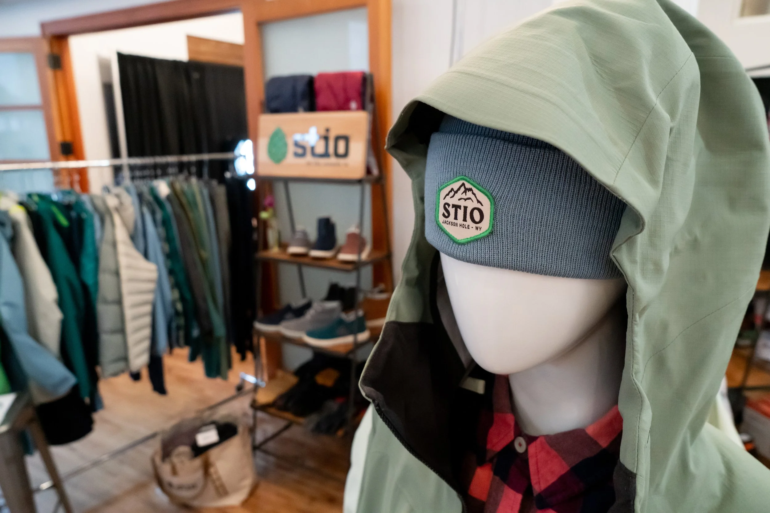 Close-up of a mannequin wearing a green jacket, a gray beanie with a Stio logo, inside a clothing store with outdoor apparel and shoes visible in the background.