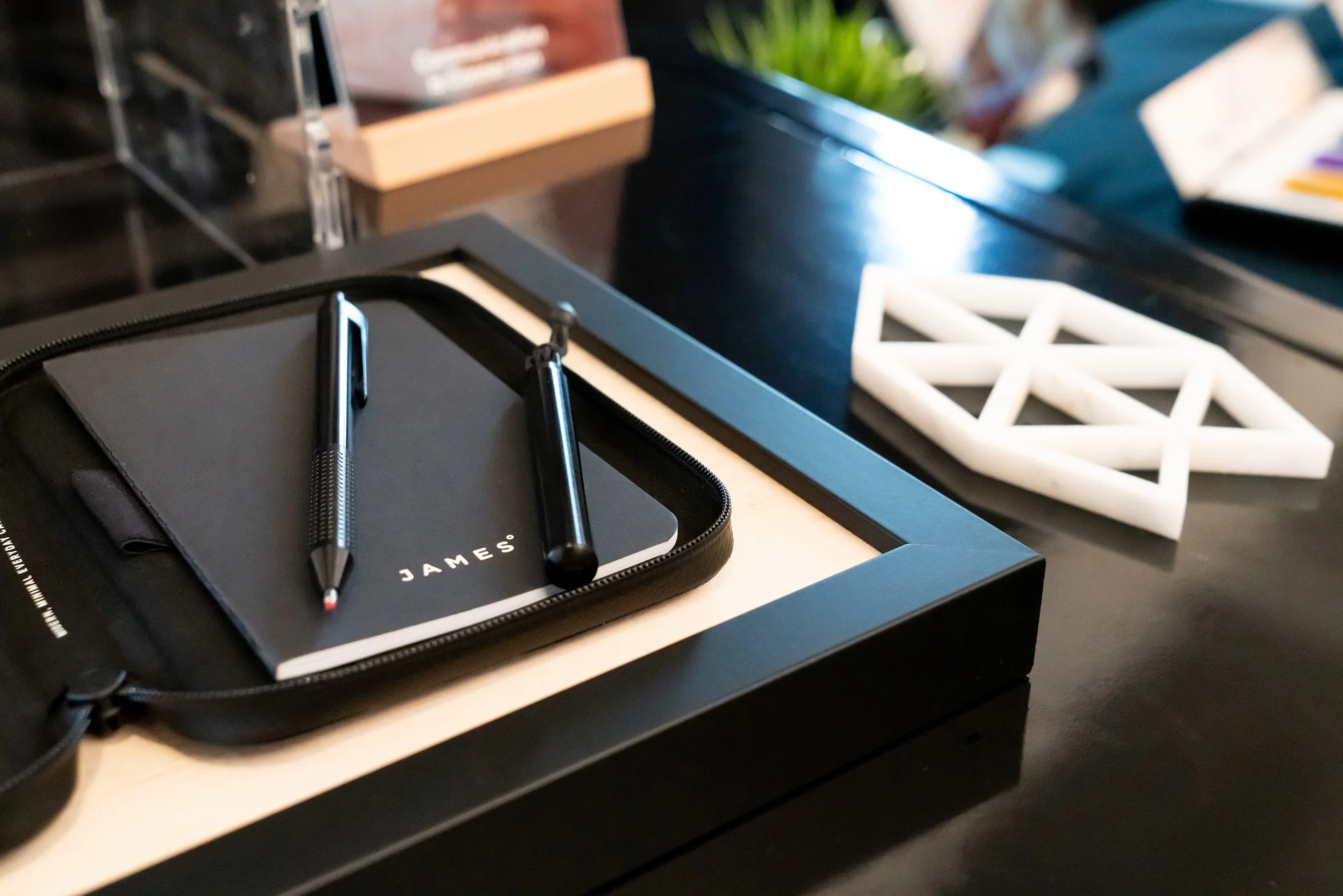 A black tray holding a black notebook with the word 'JAMES' on the cover, a black pen, and a black stylus. To the side, there is a white geometric sculpture on a dark surface, with part of a menu or magazine visible in the background.