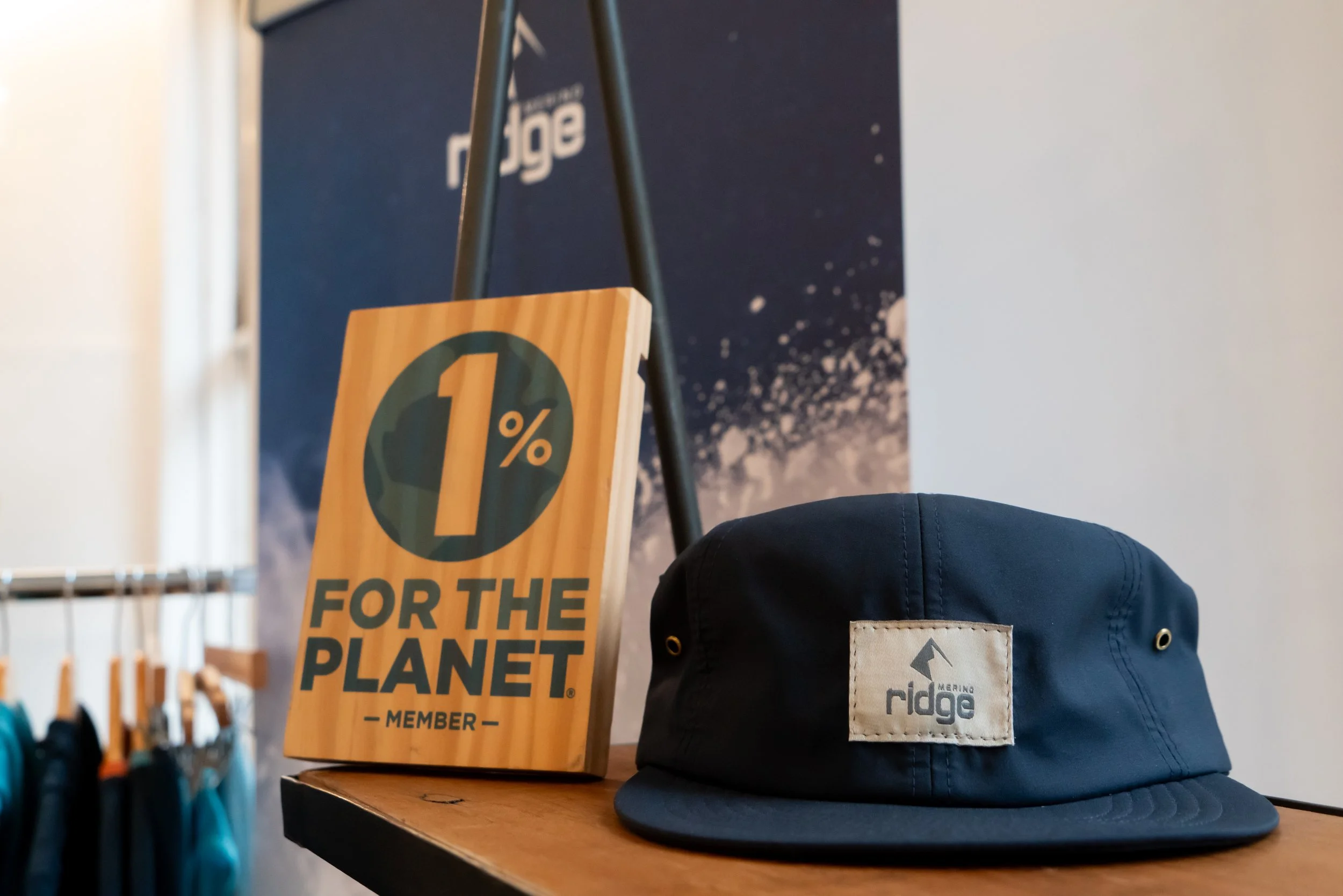 A navy blue 'ridge' cap with a white patch on the front, a Bangor logo, placed on a wooden surface. Next to the cap, there is a wooden sign with the message '1% for the Planet, Member', leaning against a booster's surface, with a dark blue background