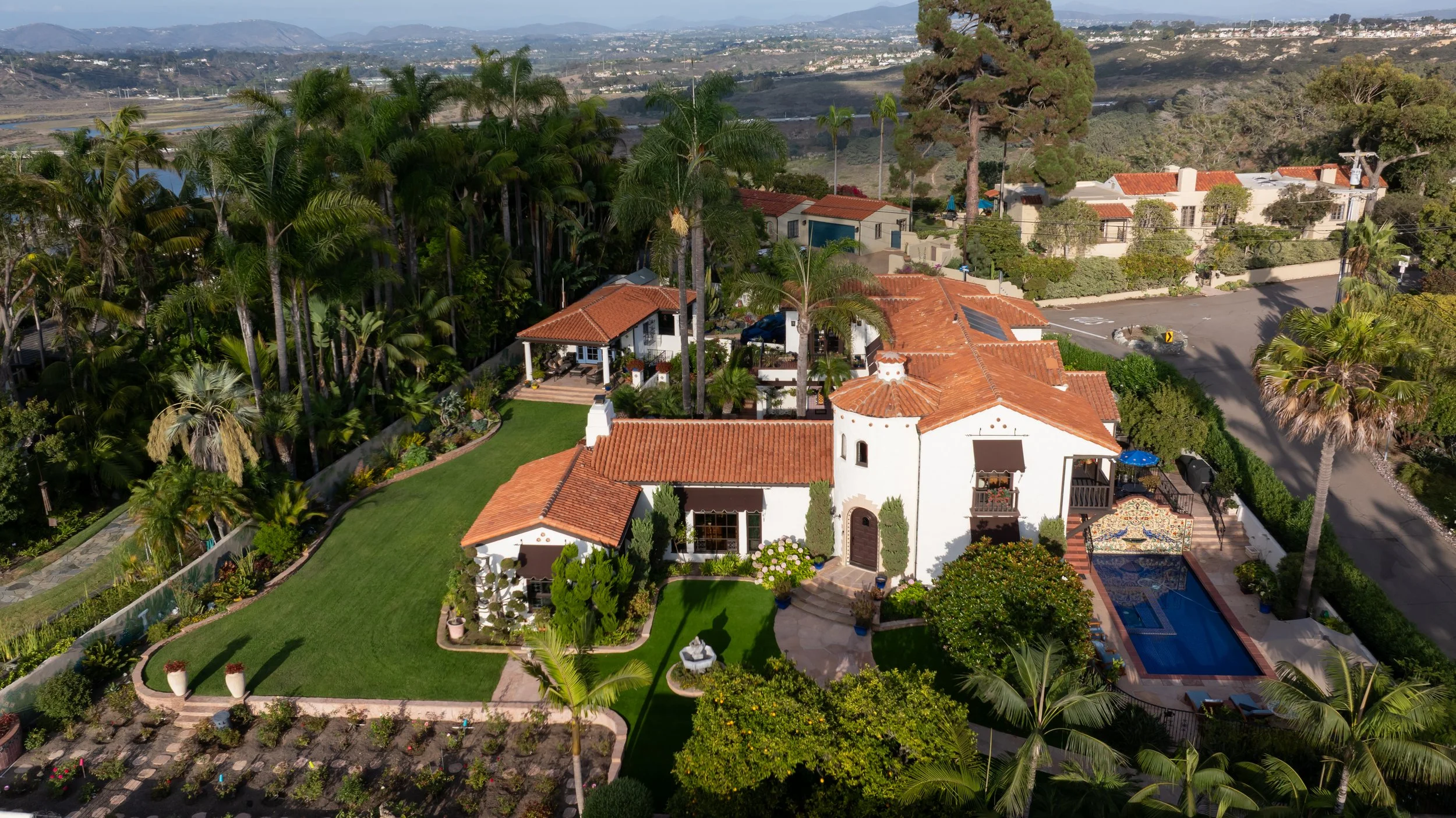 A large Mediterranean-style house with red tile roof, surrounded by lush tropical landscaping and a well-maintained lawn. Features a swimming pool with a decorative wall and patio area, and is located in a scenic area with mountains in the distance.