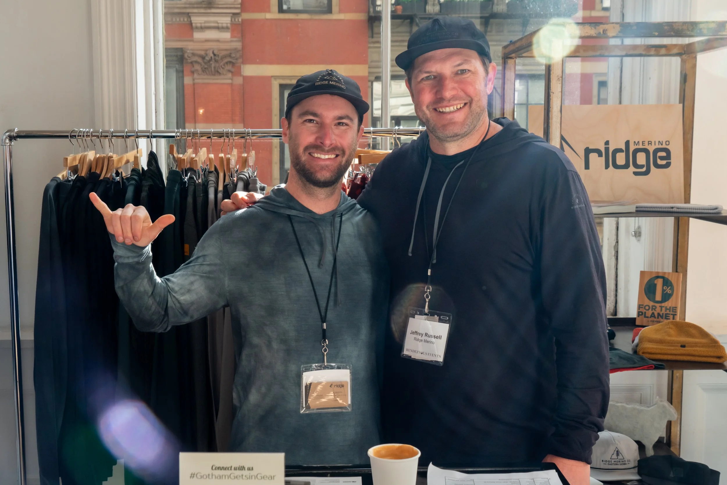 Two smiling men posing at a merchandise booth with clothing and accessories, one making a shaka sign.