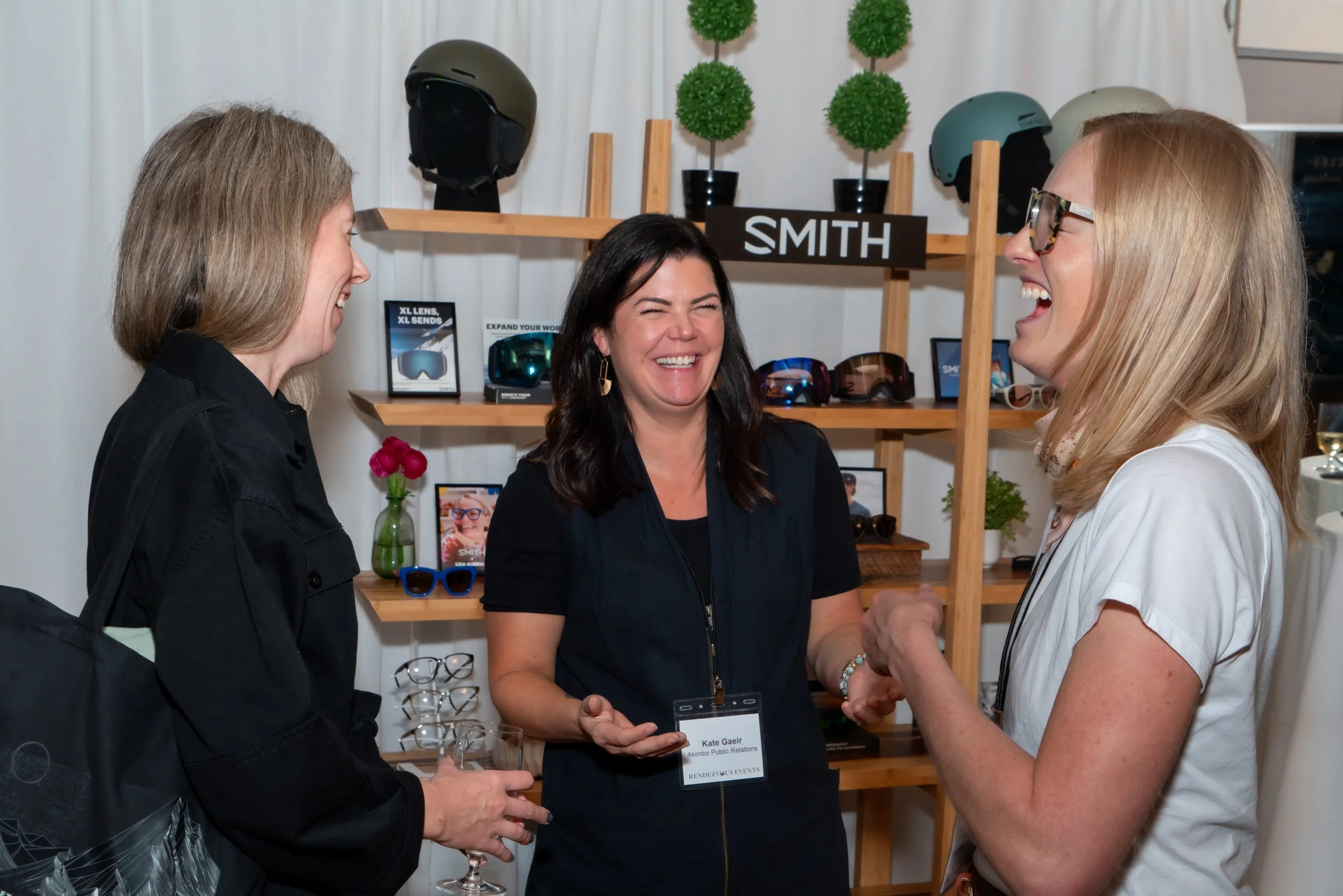 Three women are talking and laughing at a booth with sunglasses and helmets displayed on shelves behind them.