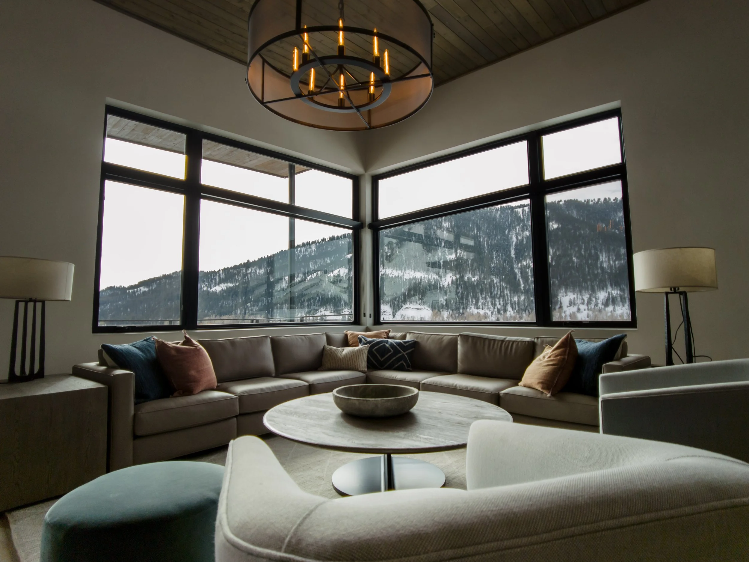 Living room with large windows showing mountain view, beige sectional sofa with pillows, round coffee table with decorative bowl, floor lamps, and a ceiling chandelier.