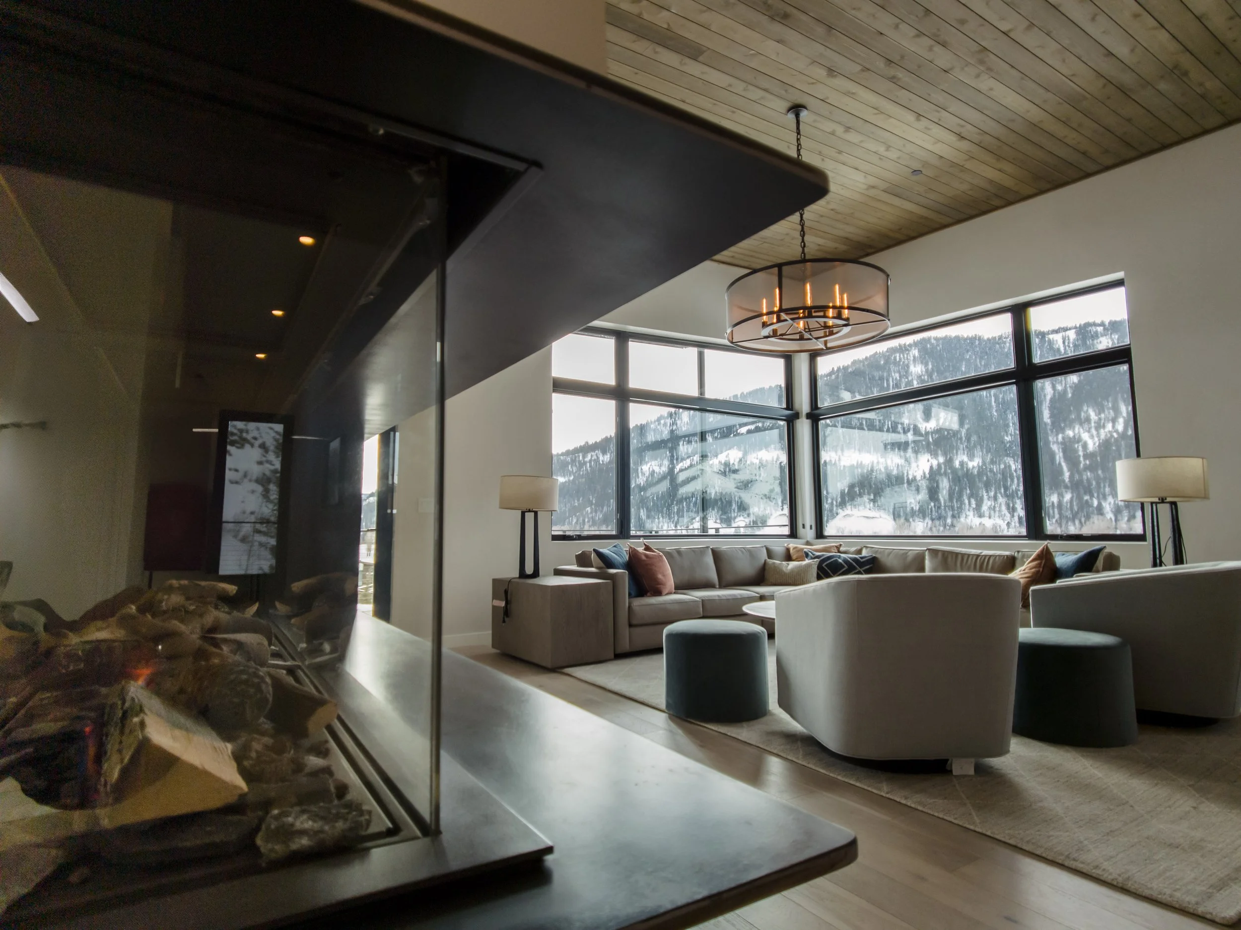 A spacious living room with large windows showing snow-covered mountains outside. The room has a sectional sofa and armchairs arranged around a central coffee table, with floor lamps on either side and a modern chandelier hanging from the wooden ceil