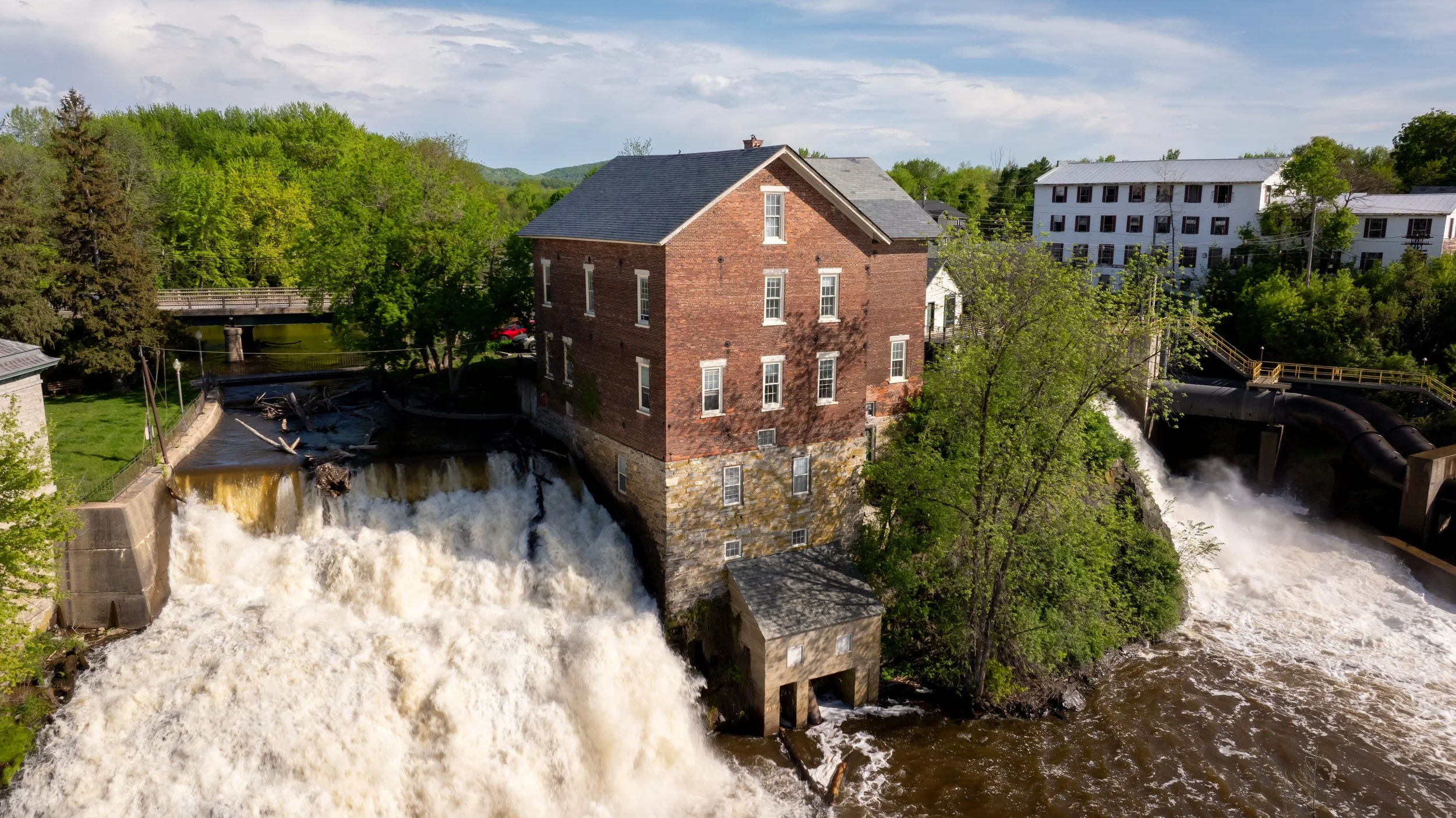 An old brick building situated next to a rushing water dam, with a small structure beneath it, surrounded by lush green trees and modern buildings in the background.