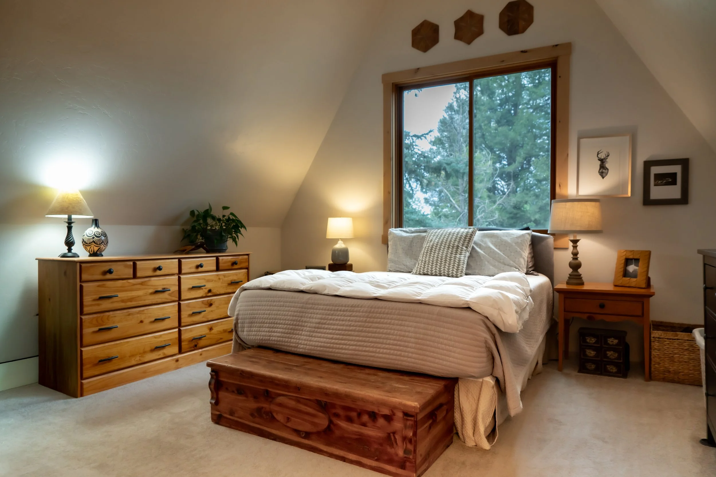 Cozy bedroom with a bed, wooden dresser, nightstand, and window showing trees outside.
