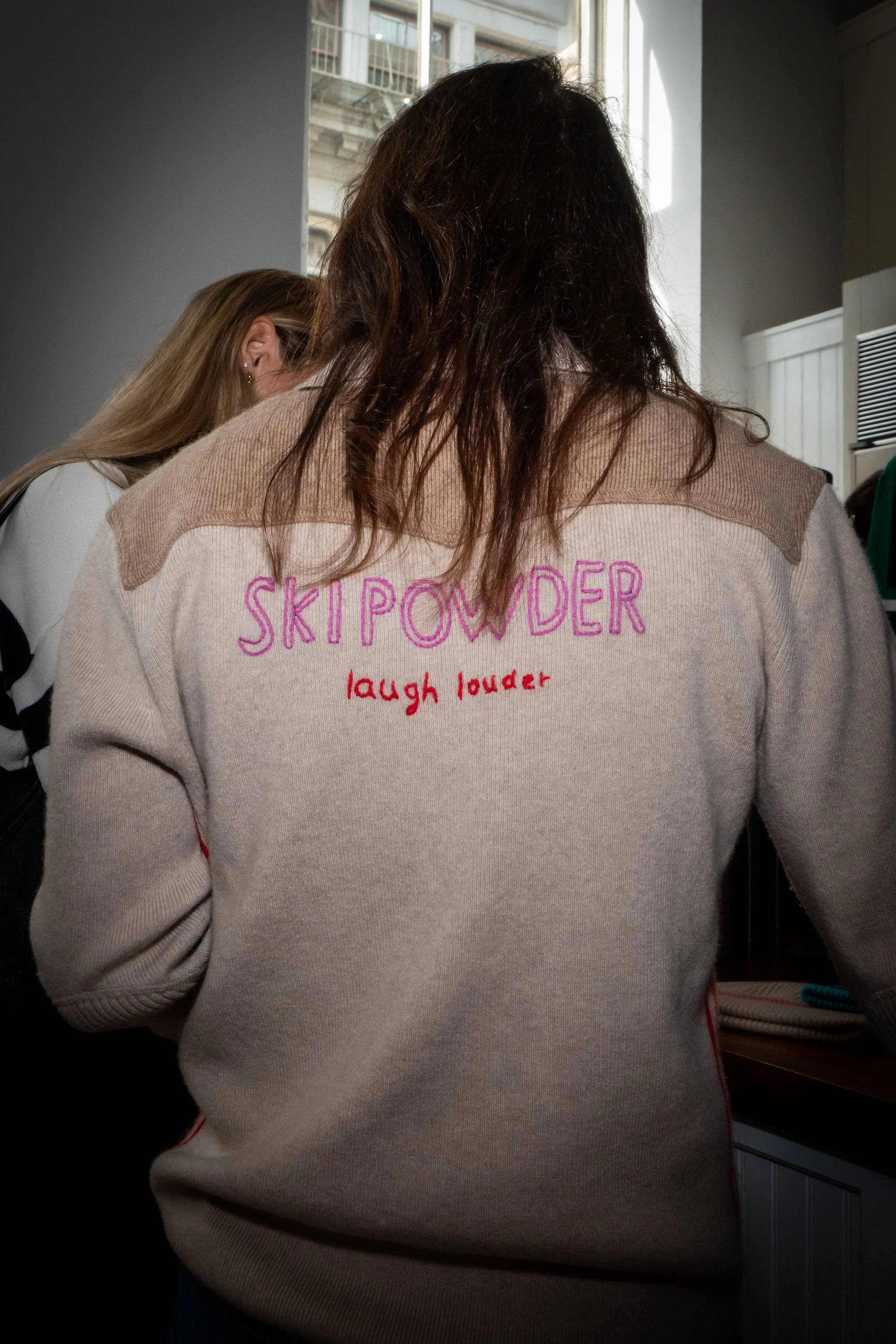 Back of a person wearing a beige sweater with the words "SKIP POWDER" embroidered in pink and "laugh louder" embroidered in red on the back. The person has long, wavy brown hair.