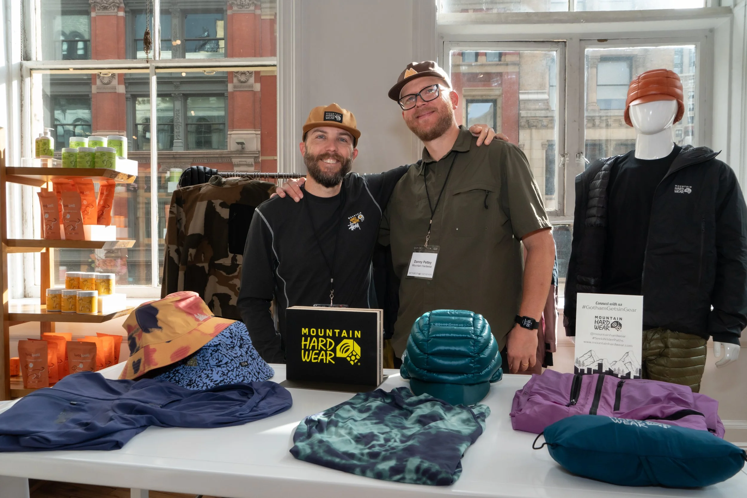 Two men standing behind a table with outdoor gear and clothing, in a brightly lit room with large windows, one man in a black shirt and the other in a green shirt, both smiling, with a mannequin wearing a black jacket and an orange hat to their right