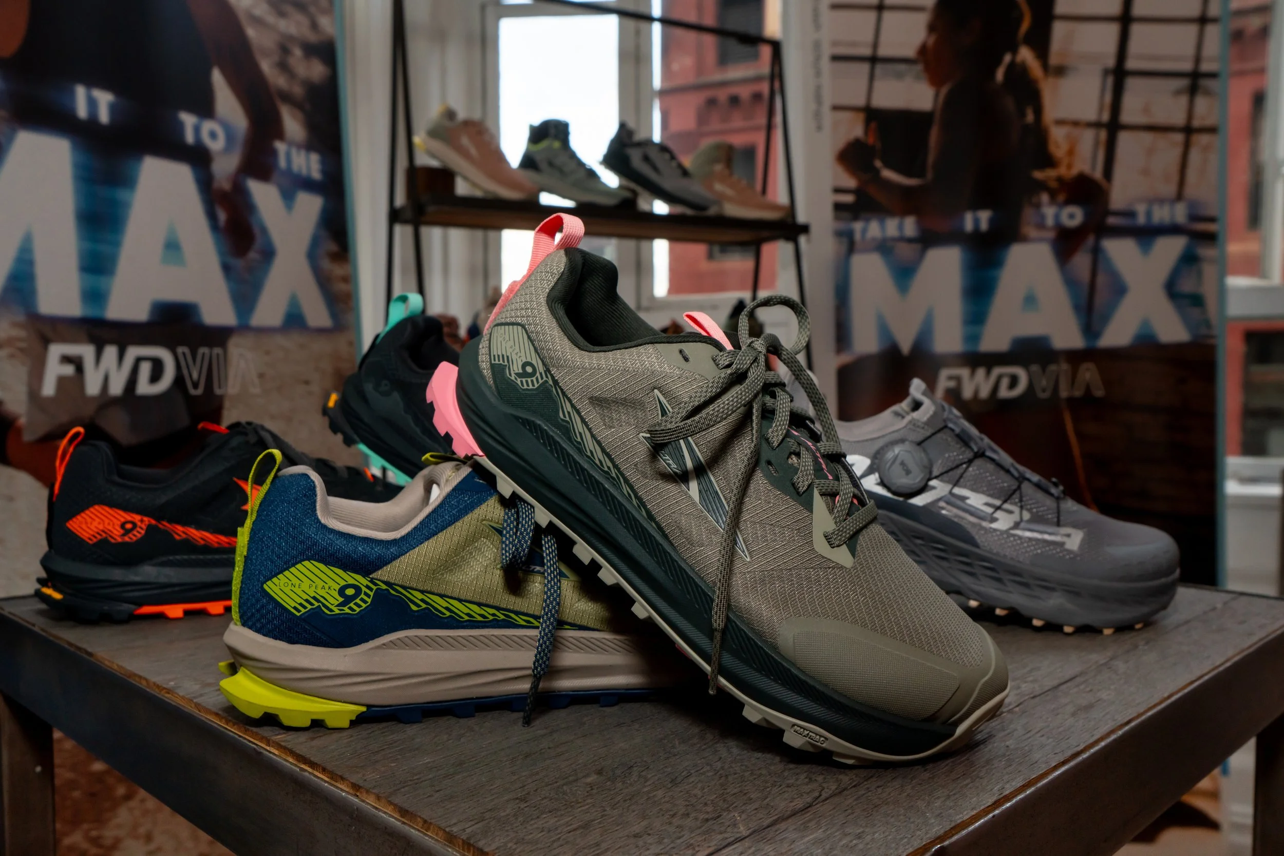 A display of trail running shoes on a wooden table, with two shelves of additional shoes in the background, inside a store with large windows showing a brick building outside.