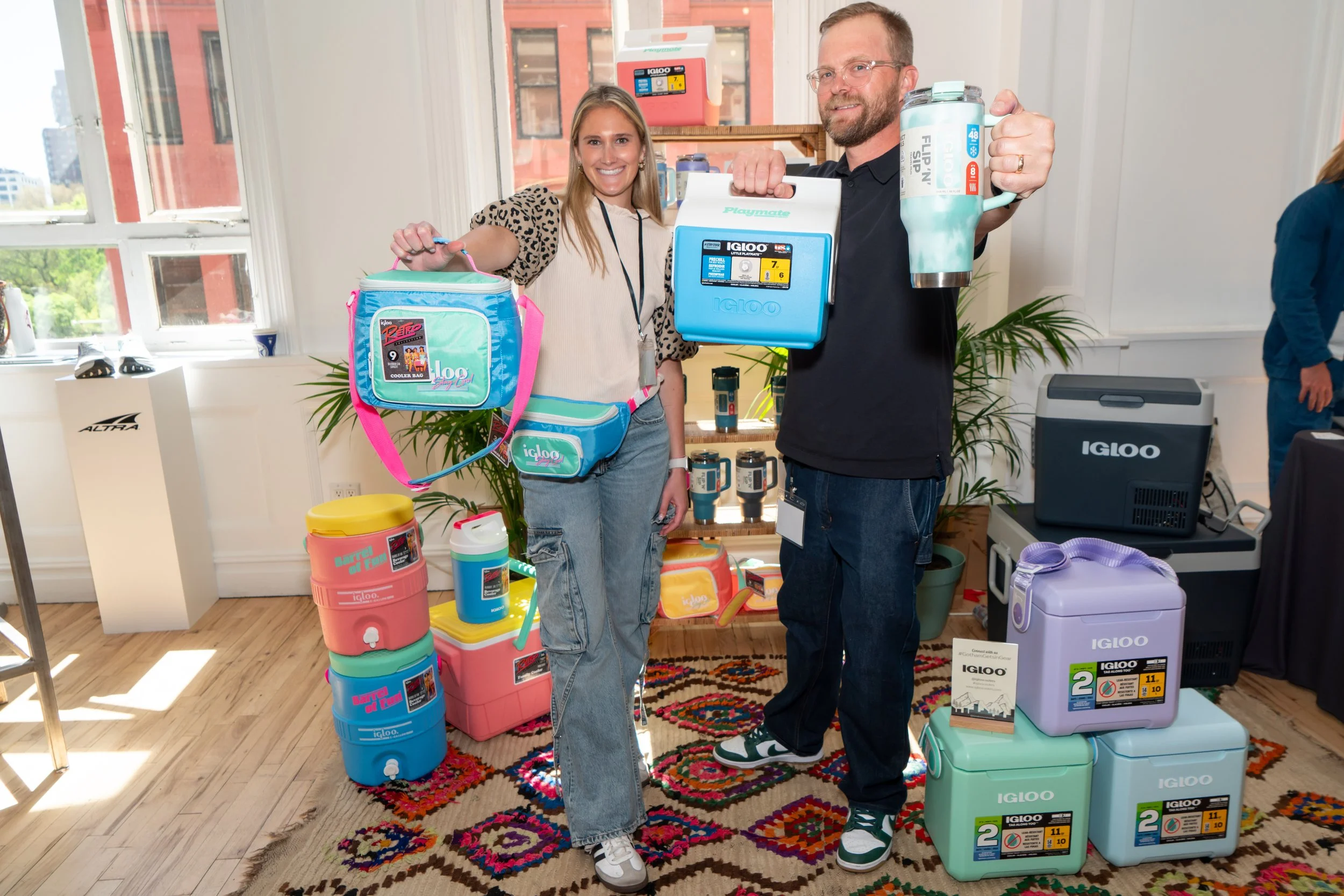 Two people smiling at a trade show booth displaying Igloo coolers and other insulated containers, with a woman holding a pink and blue cooler bag and a man holding a large turquoise and white insulated tumbler and a blue mini fridge.