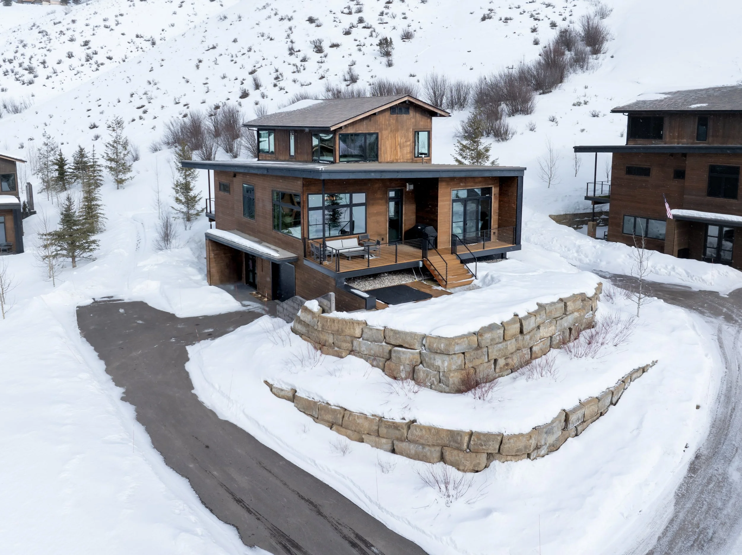 Modern multi-level house with wooden exterior and large windows, situated in a snowy mountainous landscape, with a stone retaining wall and a winding driveway in the foreground.