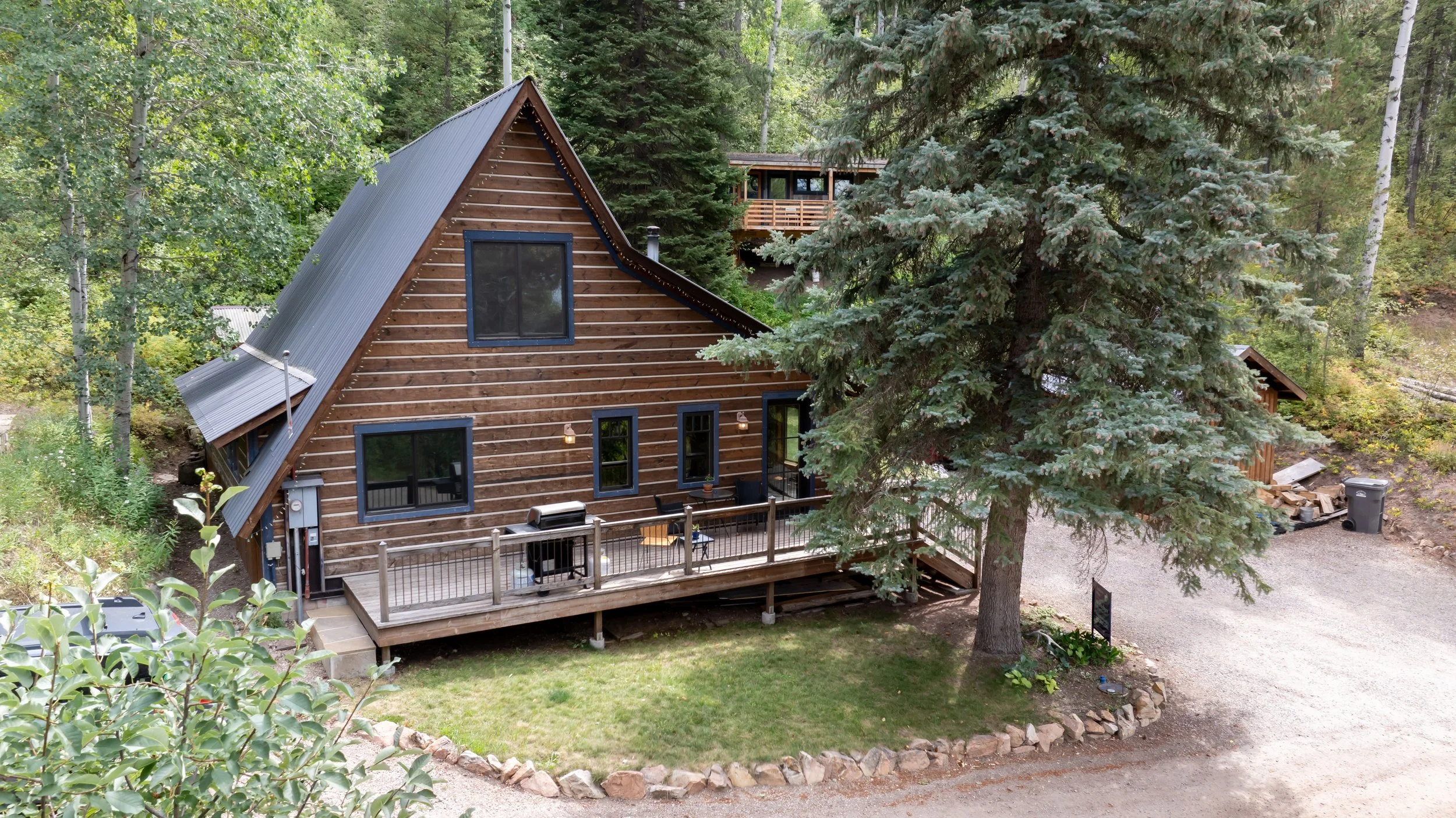 A rustic wooden house with a steep metal roof, large windows, and a deck with outdoor furniture, situated in a wooded area with trees and a gravel driveway.