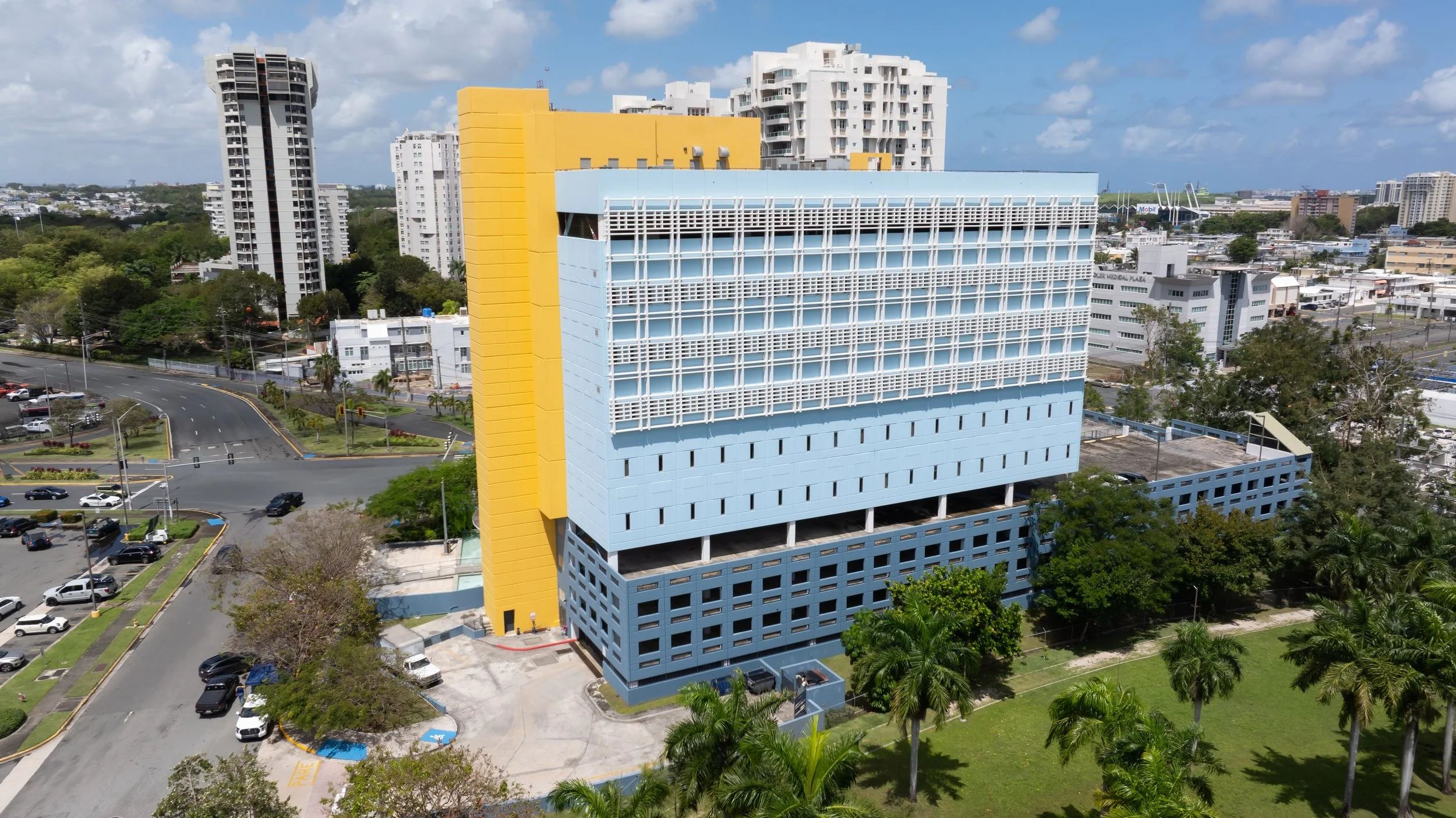 Aerial view of a colorful multi-story building with a blue facade, yellow accent wall, and white window panels, surrounded by trees, parking lots, and cityscape with high-rise buildings under a partly cloudy sky.