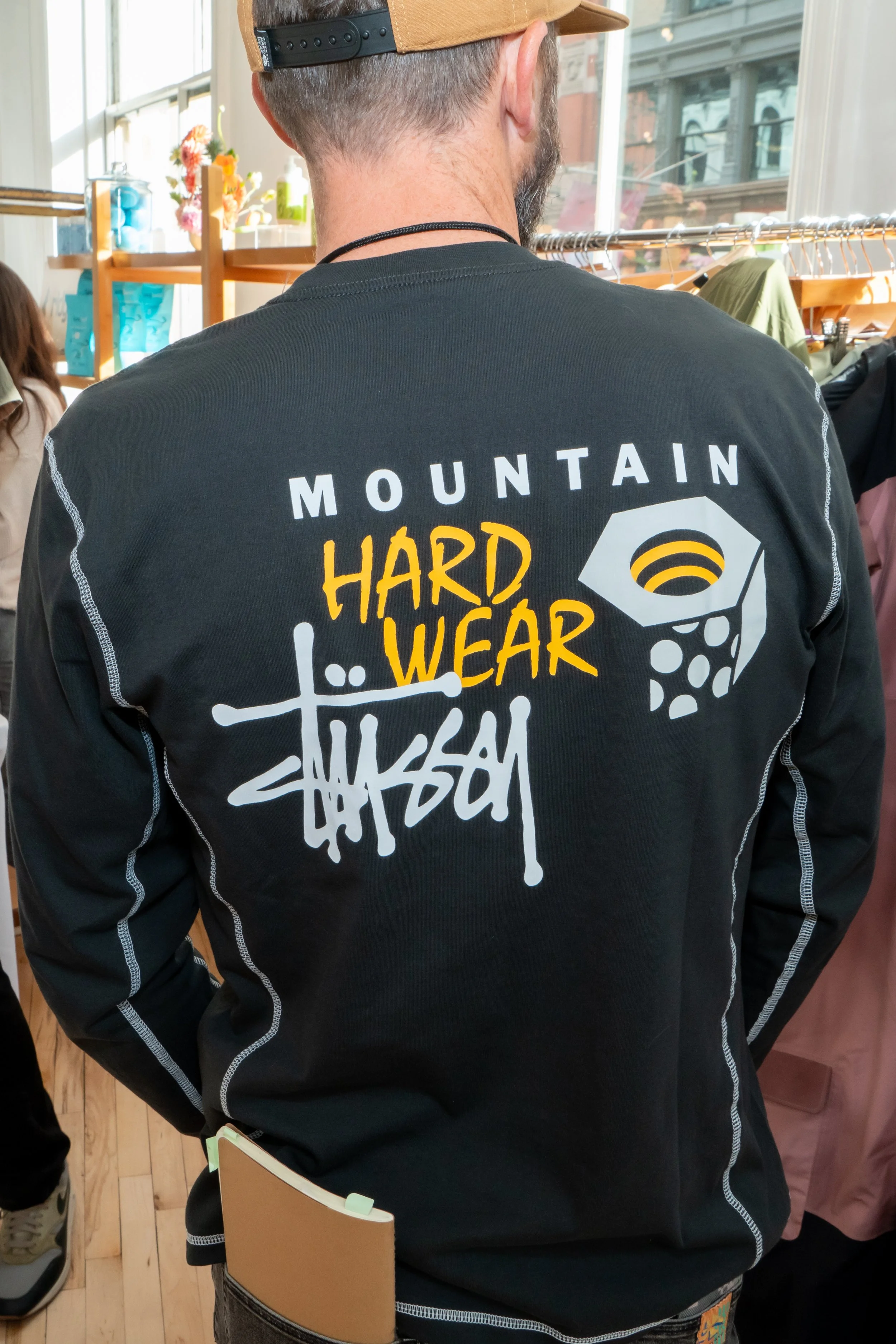 Person wearing a black jacket with white and yellow text and graphics, including the phrase 'MOUNTAIN HARD WEAR' and a dice graphic, inside a store with clothing racks and large windows.