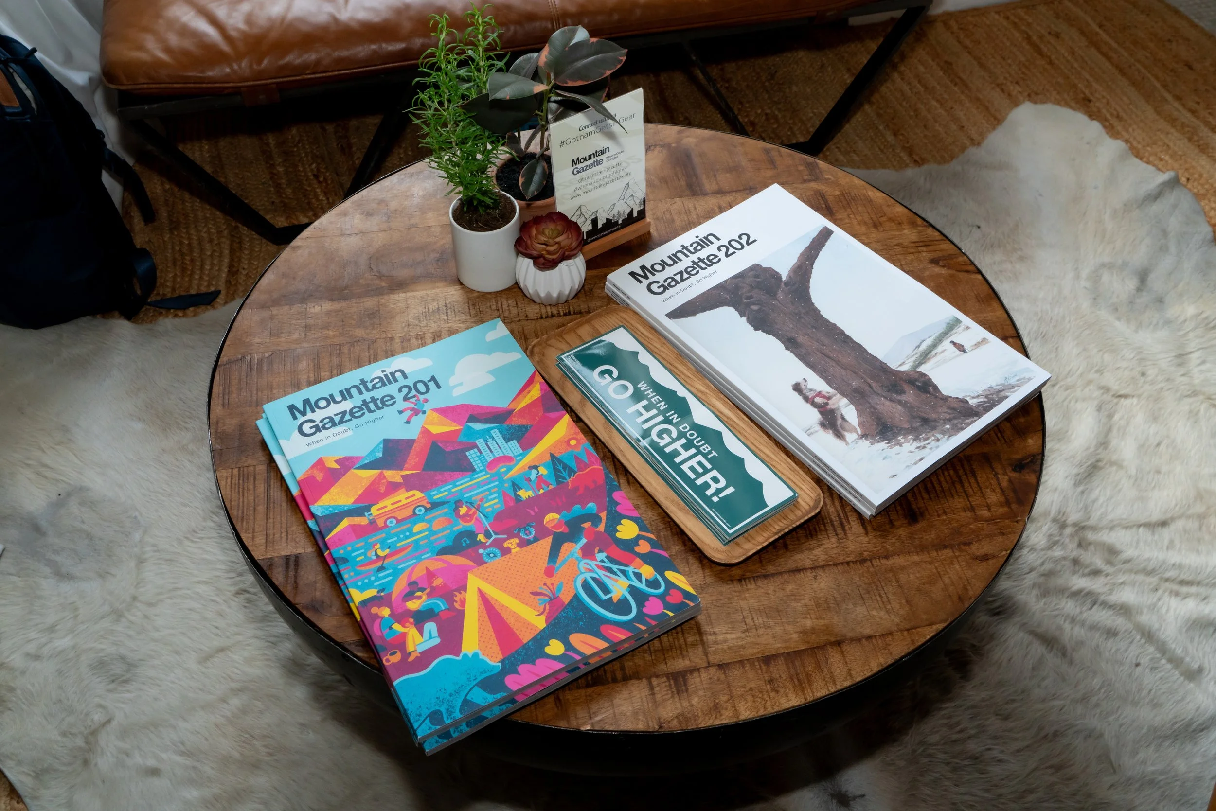 Wooden table with magazines, potted plants, and a sign, surrounded by fur rugs.