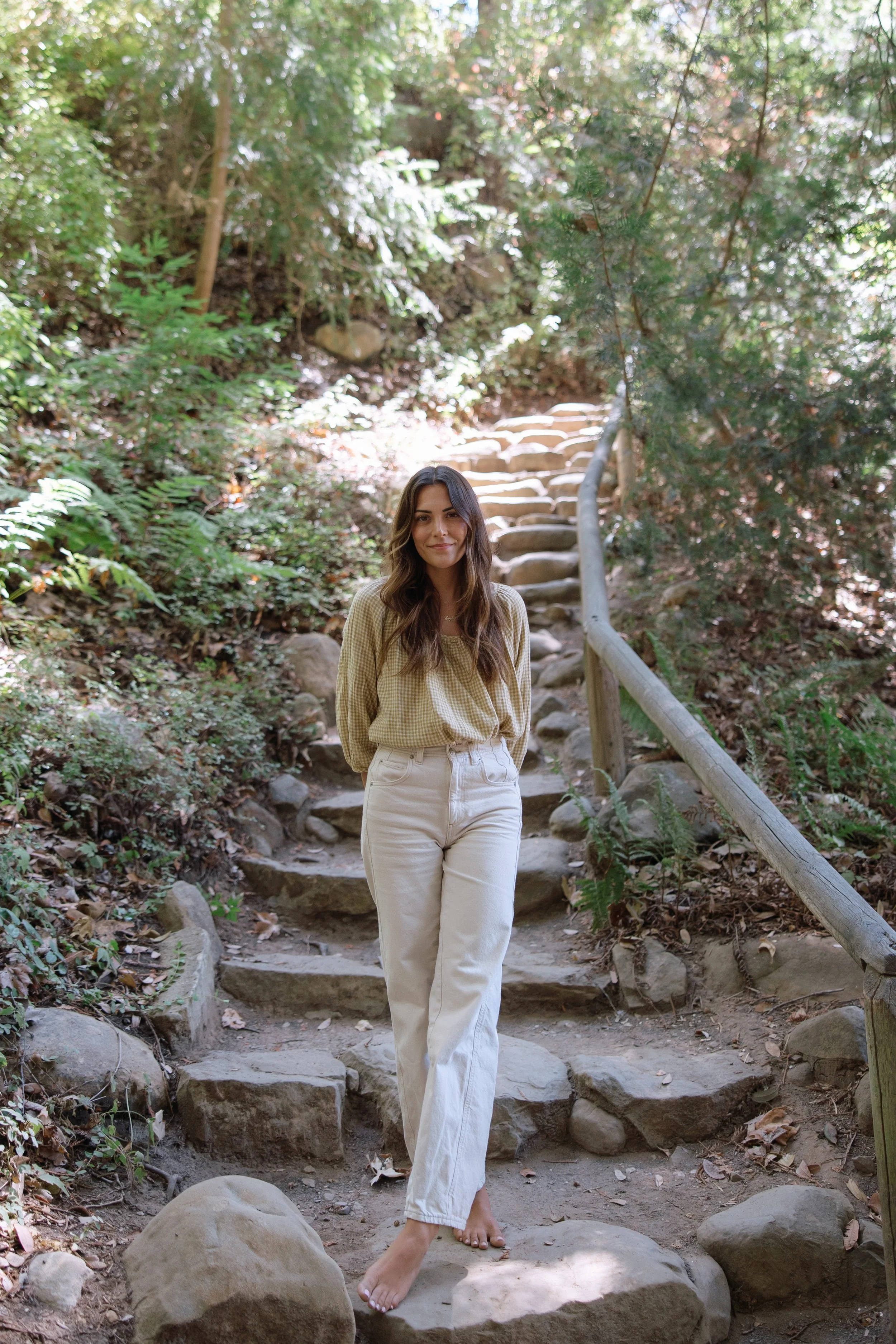 A woman with long wavy brown hair wearing a yellow-green checkered shirt and white jeans walking barefoot on a rocky trail surrounded by trees.