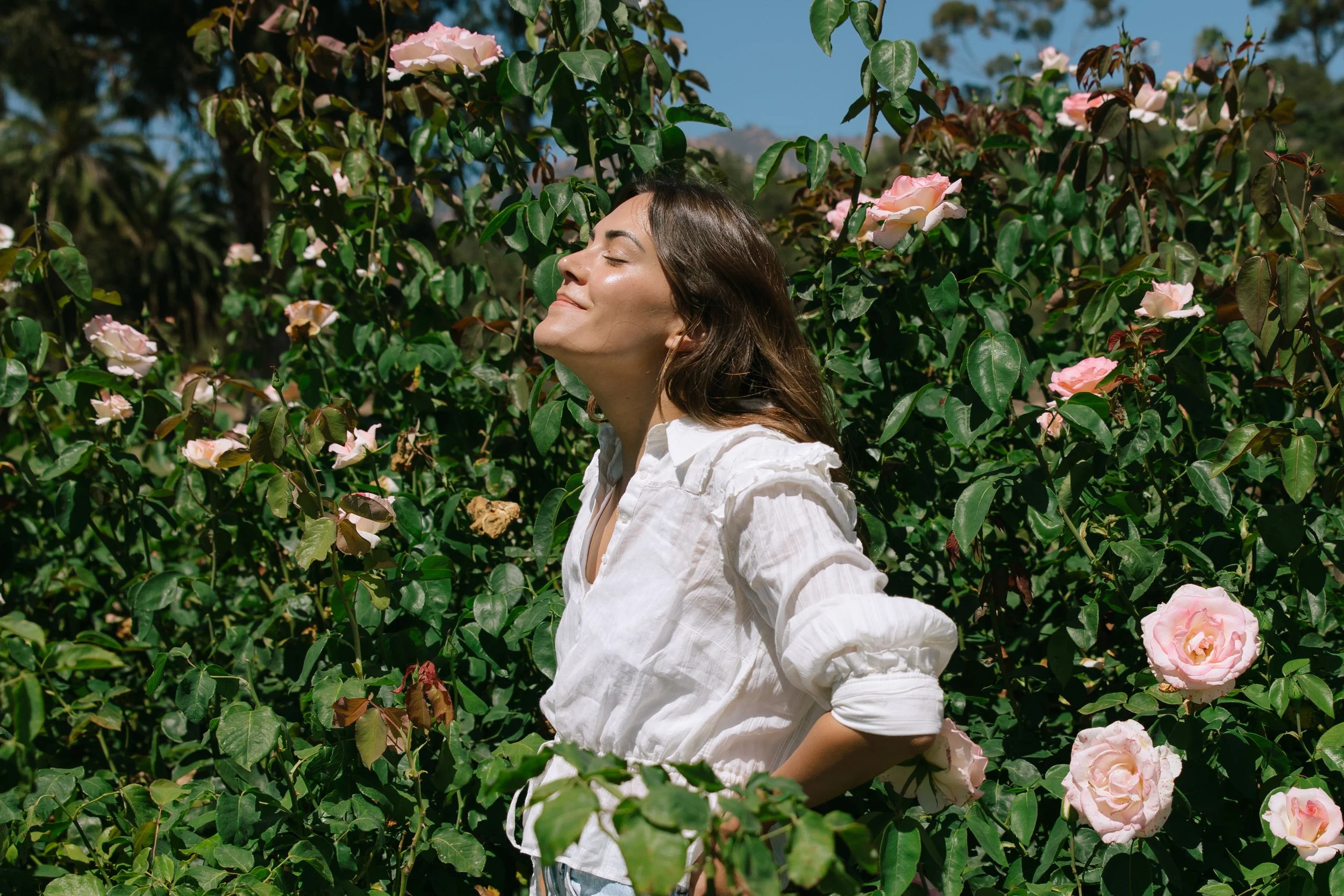 A woman standing in a rose garden with her eyes closed and a smile on her face, wearing a white shirt.