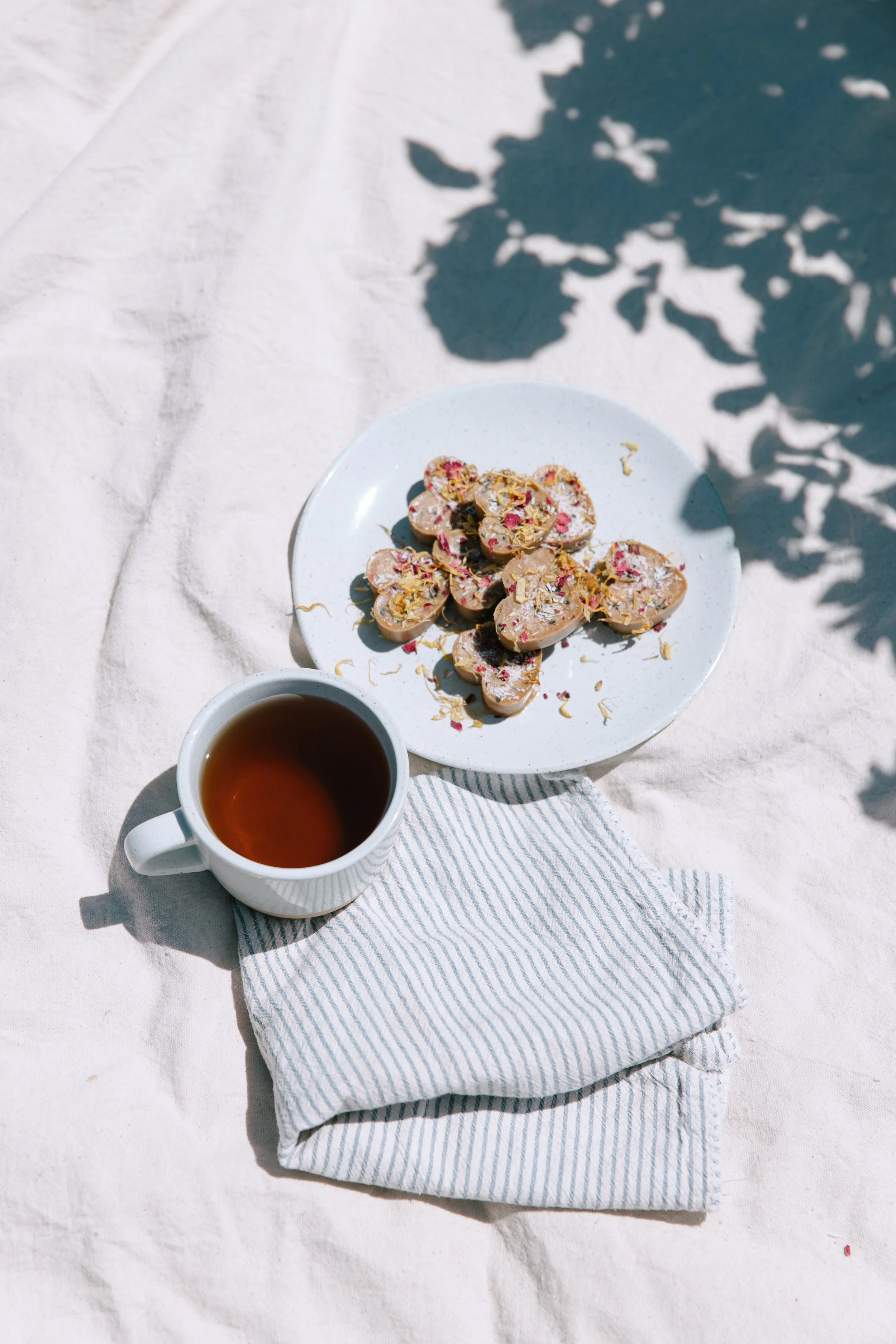 A white plate with heart-shaped cookies topped with sprinkles and flower petals, a cup of tea, and a striped cloth napkin on a light fabric surface, with shadows of leaves overhead.