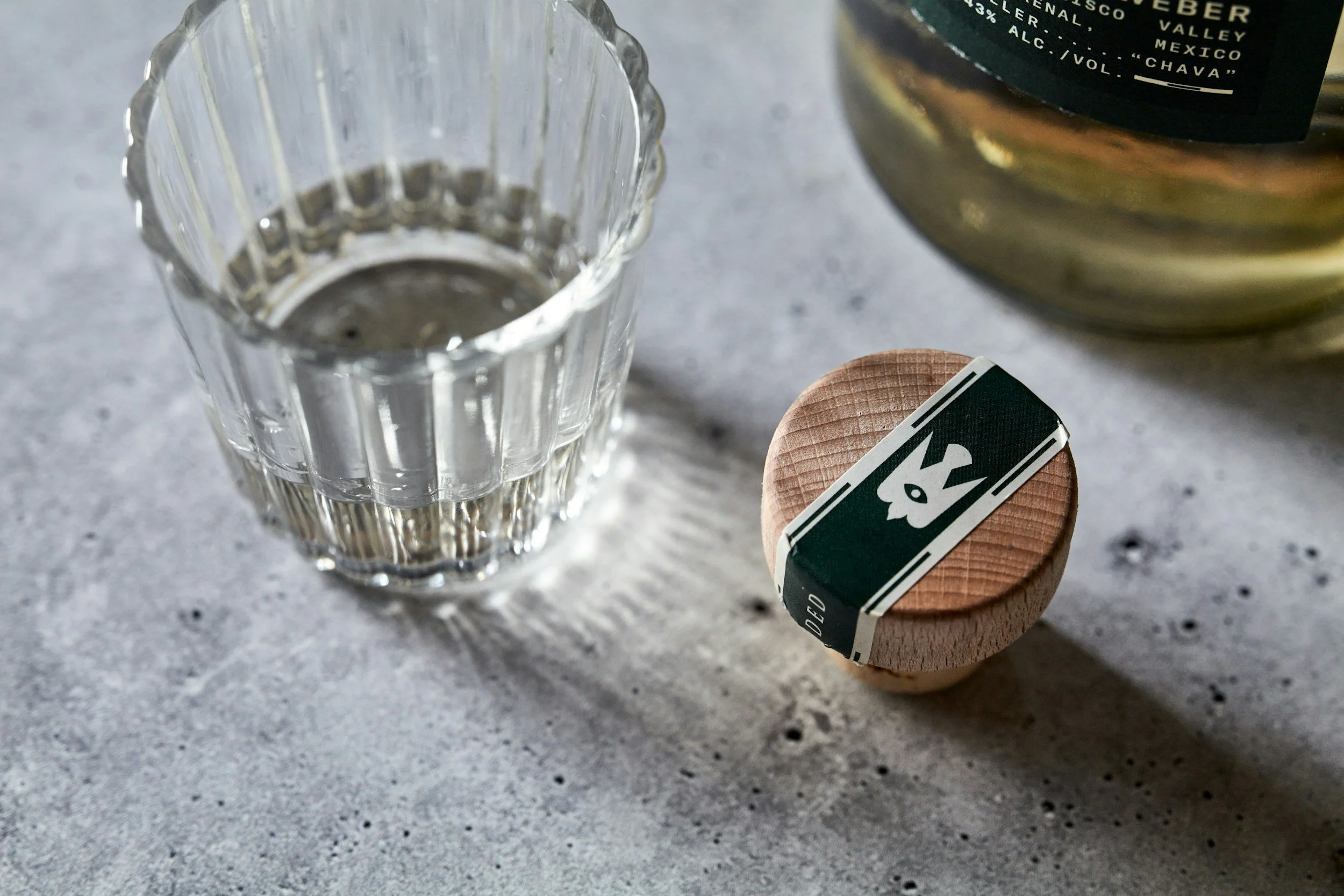 A ridged shot glass with a small amount of mezcal, next to a wooden bottle cork and the bottom of a mezcal bottle.