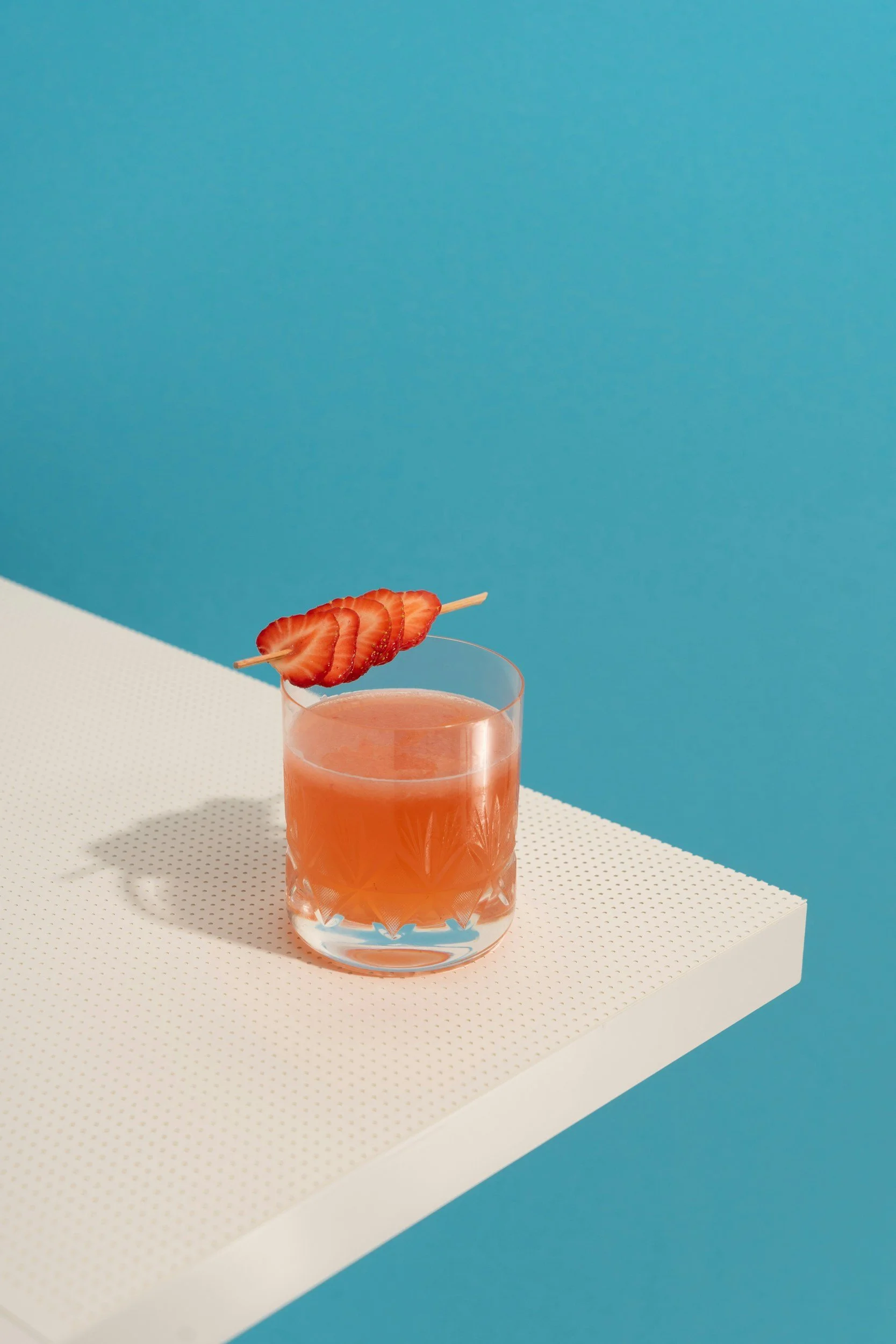 A short cocktail glass with a pinkish orange liquid, garnished with 5 strawberry slices on a skewer, sitting atop a white counter against a blue background.