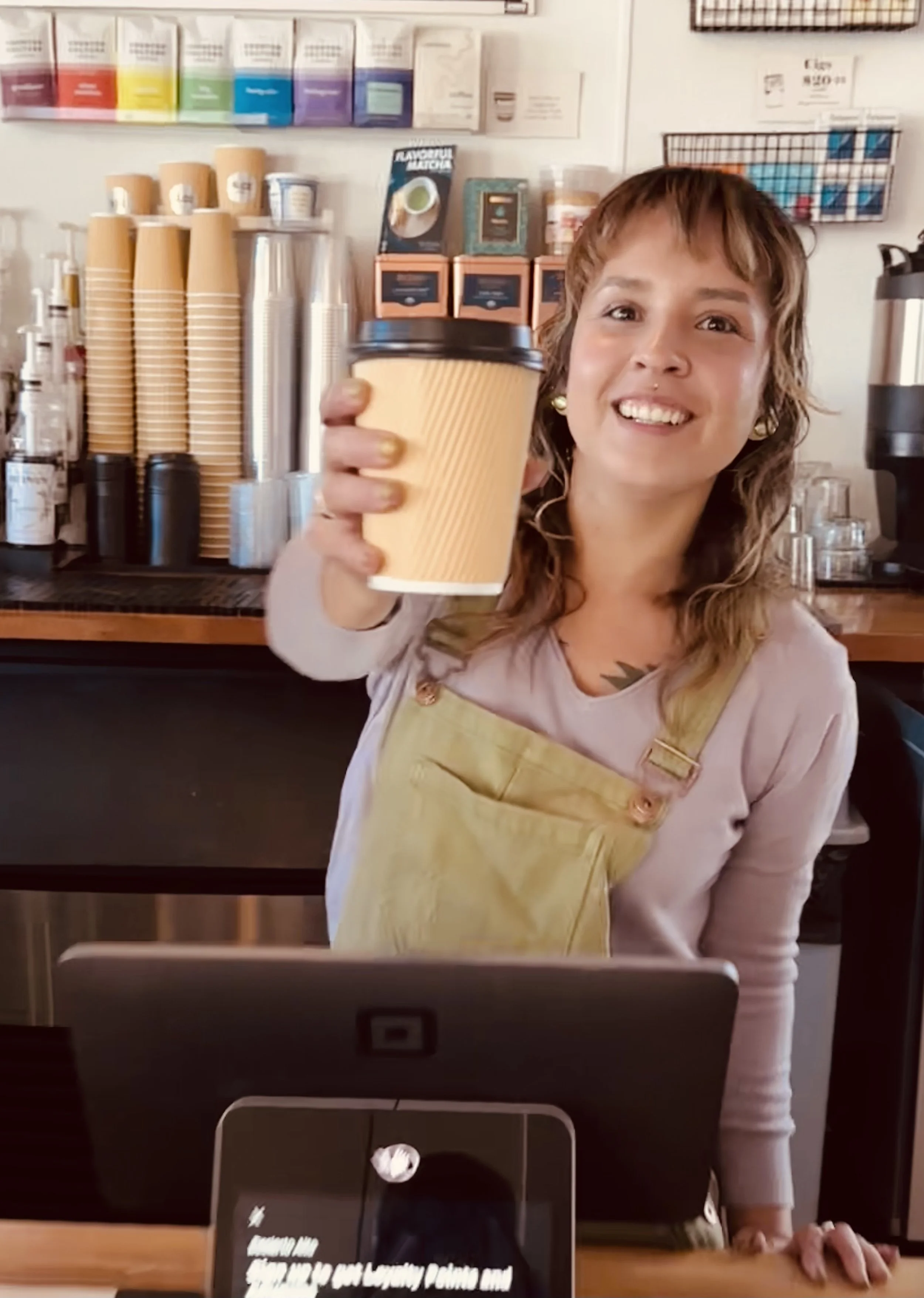 A pretty, smiling woman in faded green overalls and a lavender shirt reaches over the shop iPad to hand you a medium sized coffee cup to go. Behind her are stacks of clean coffee cups and lids, syrups, glassware, bags of coffee beans, and tea tins.