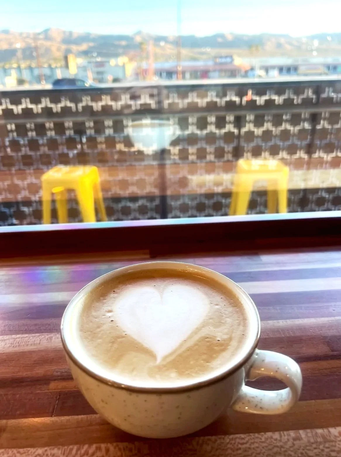 Heart-shaped latte art in a speckled mug on a wooden countertop with a windo-view of an outdoor patio with yellow high top stools.