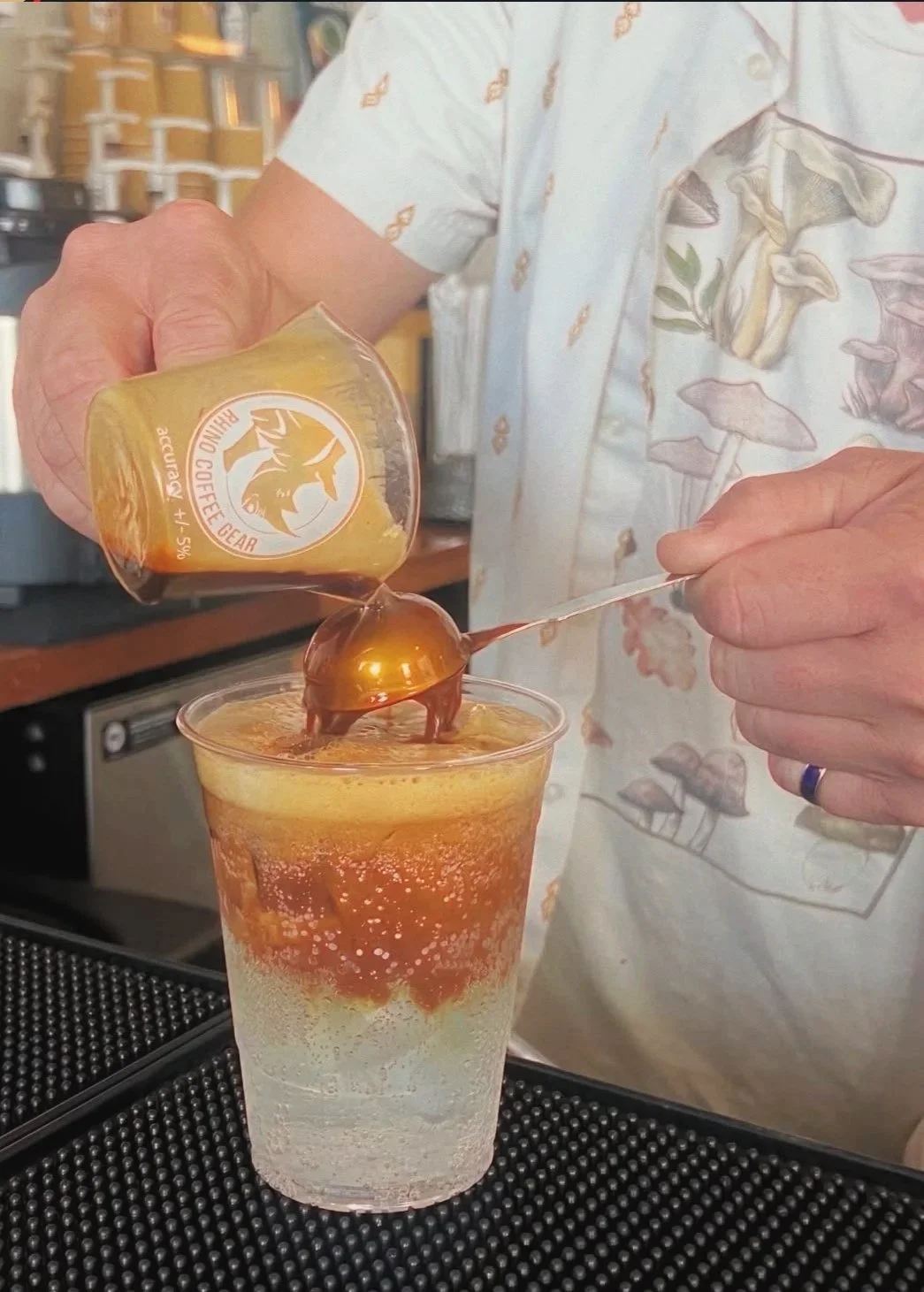 A man wearing a white button down tee makes an espresso and tonic to go, using a tablespoon to pour the espresso, which layers on top of the tonic and ice.