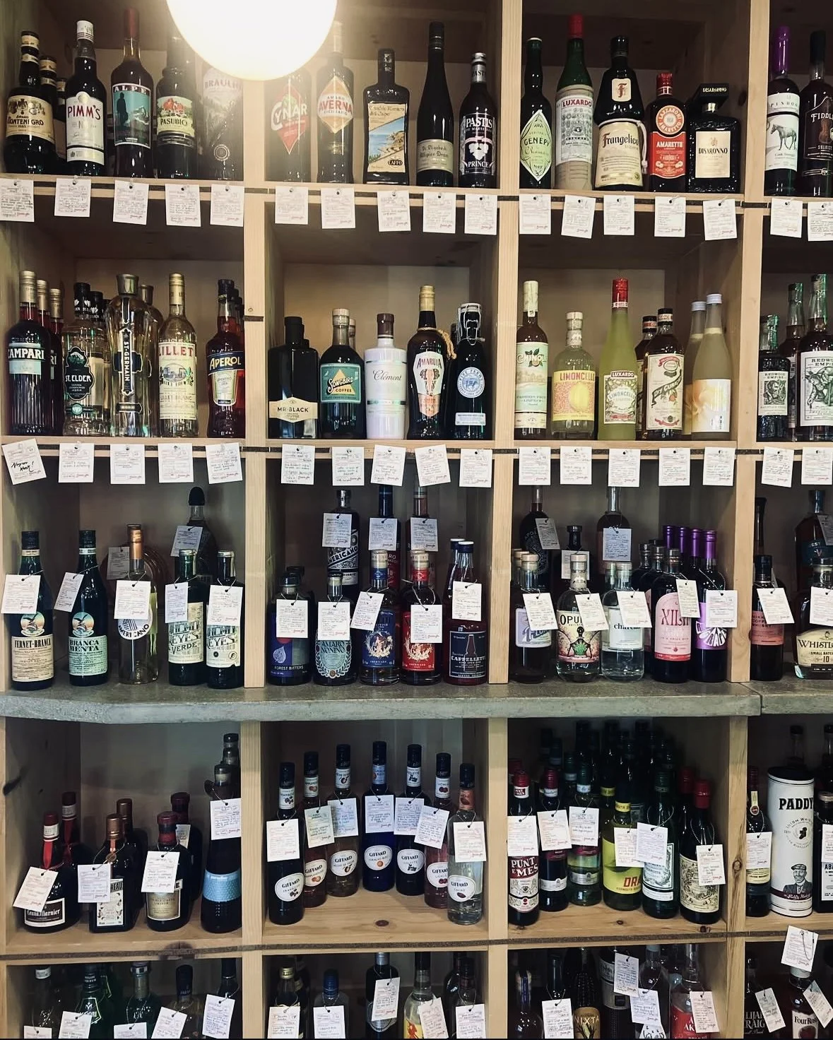 A four-tiered shop shelf stacked with about 70 liqueur and spirit bottles, all with individual description tags