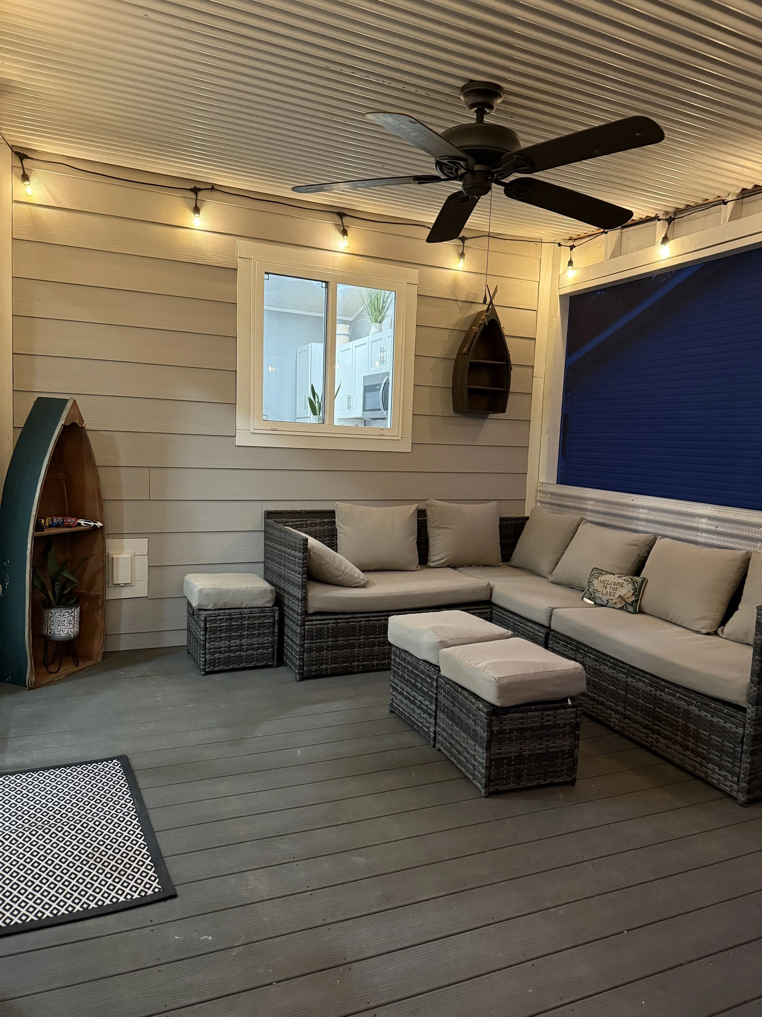 Covered outdoor patio with wicker sectional sofa with cushions, a small ottoman, a window, string lights, a ceiling fan, decorative boats hanging on wall, and a small rug.