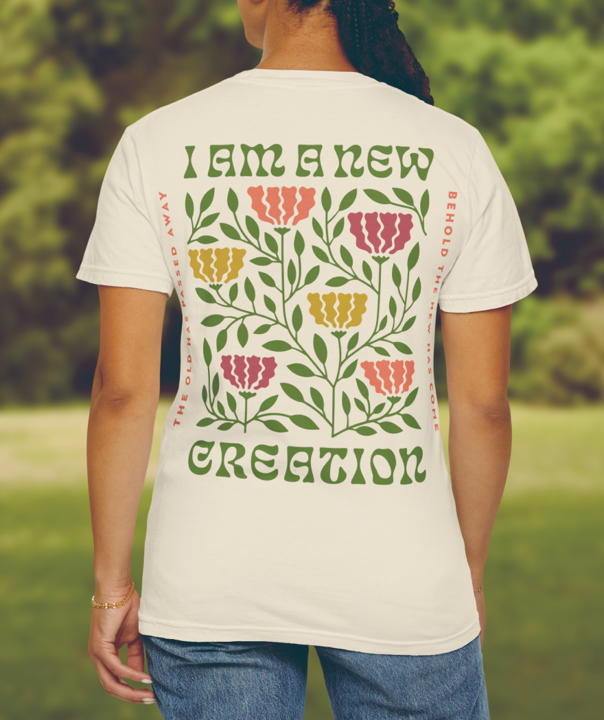 Back of a woman wearing a white T-shirt with a colorful floral graphic and text that reads 'I am a new creation' and 'Behold the new has come'.