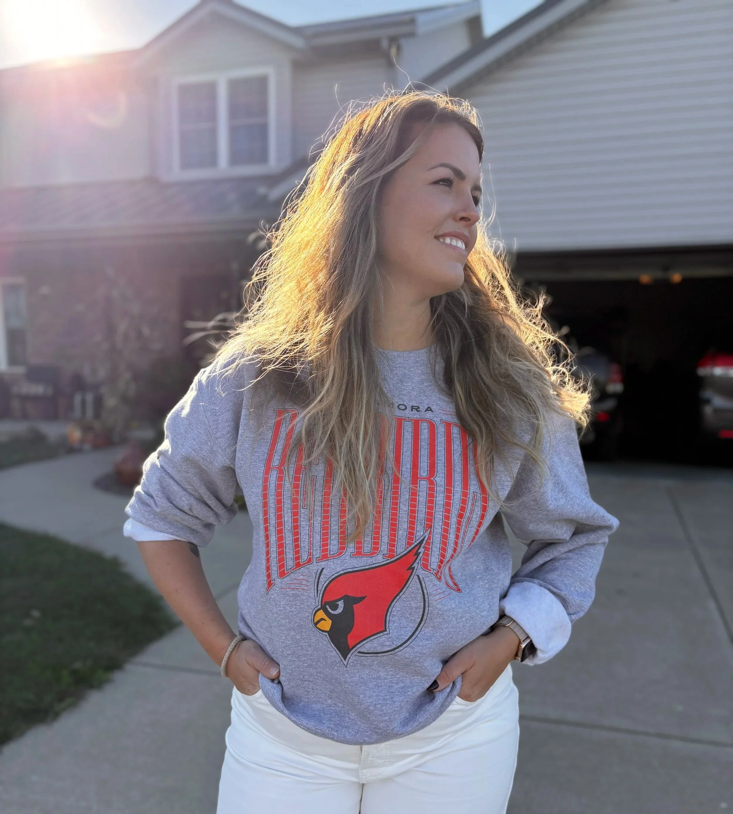 A woman with long, wavy blonde hair standing outside in front of a house, wearing a gray sweatshirt with the St. Louis Cardinals logo, smiling and looking to her right.