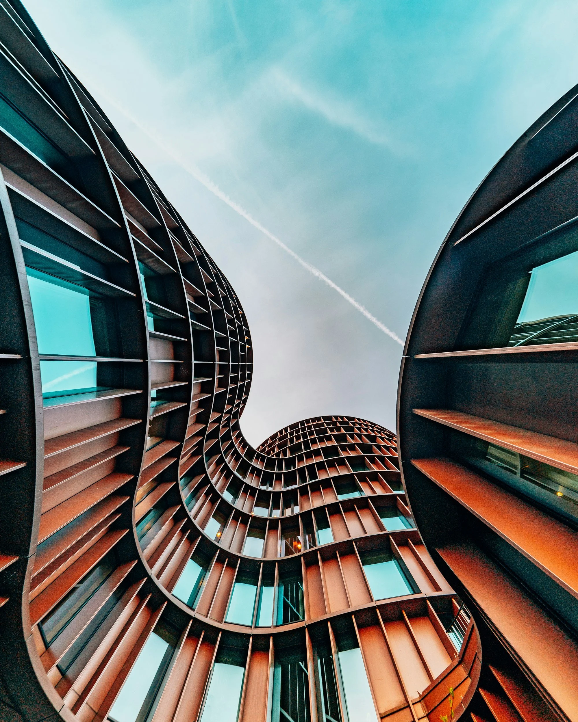 Low-angle view of a modern, curved building with glass windows and metal framework against a blue sky with a contrail.