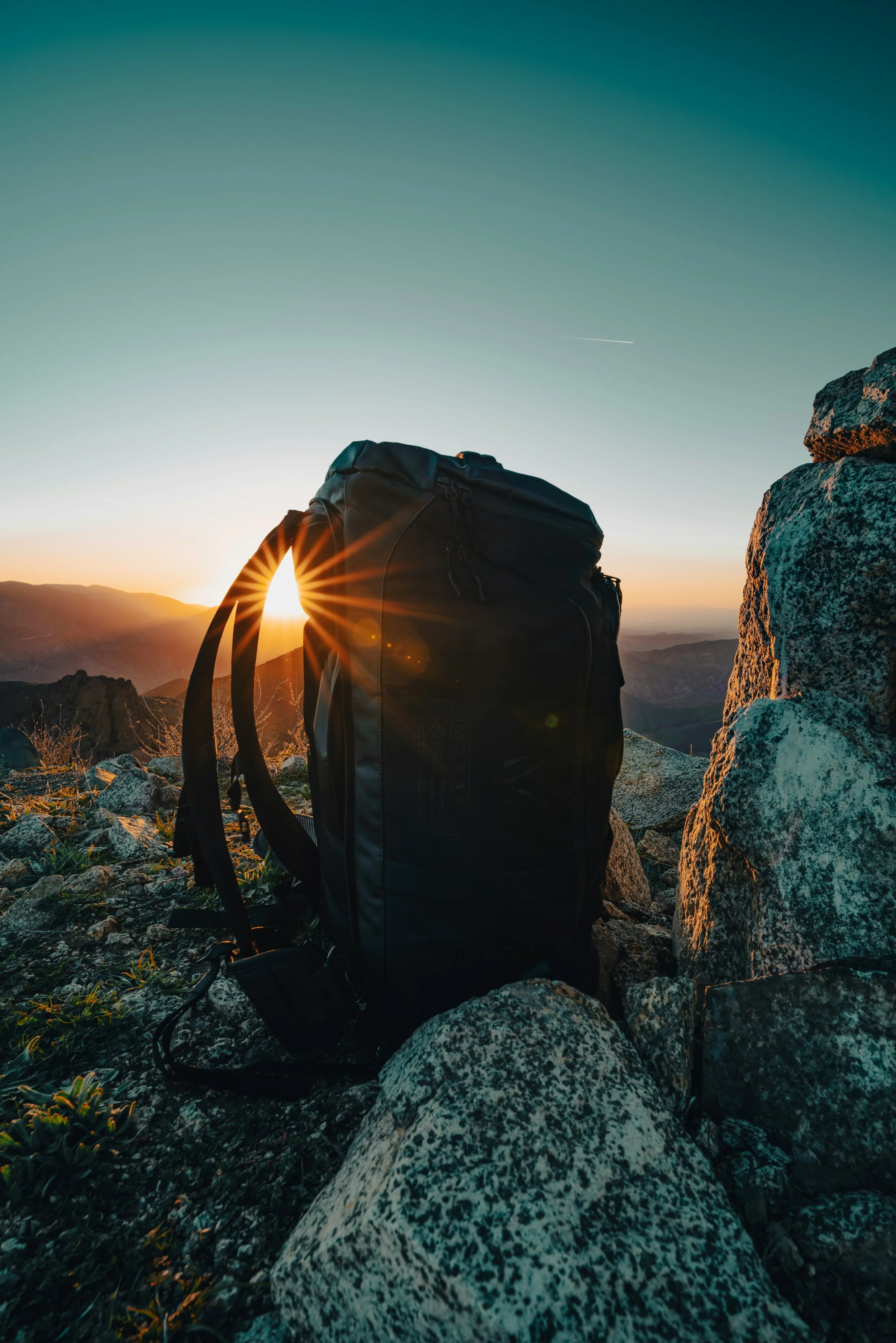 A black hiking backpack on rocky terrain during sunset with mountains in the background and a clear sky.