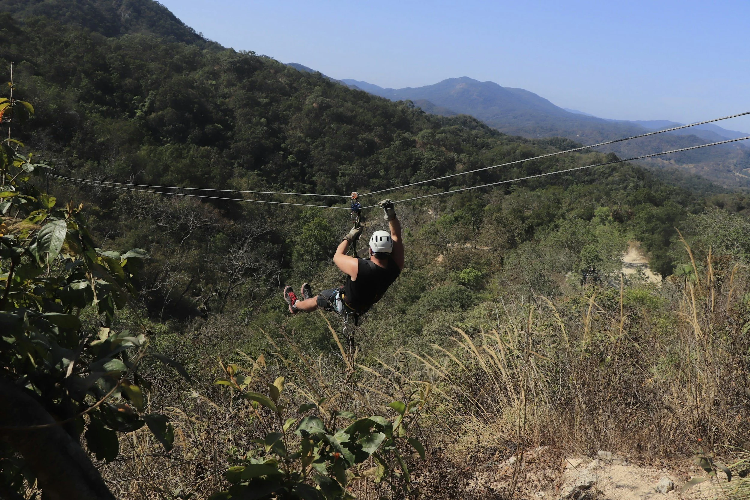 A person zip-lining above a forested mountain landscape with clear blue sky.