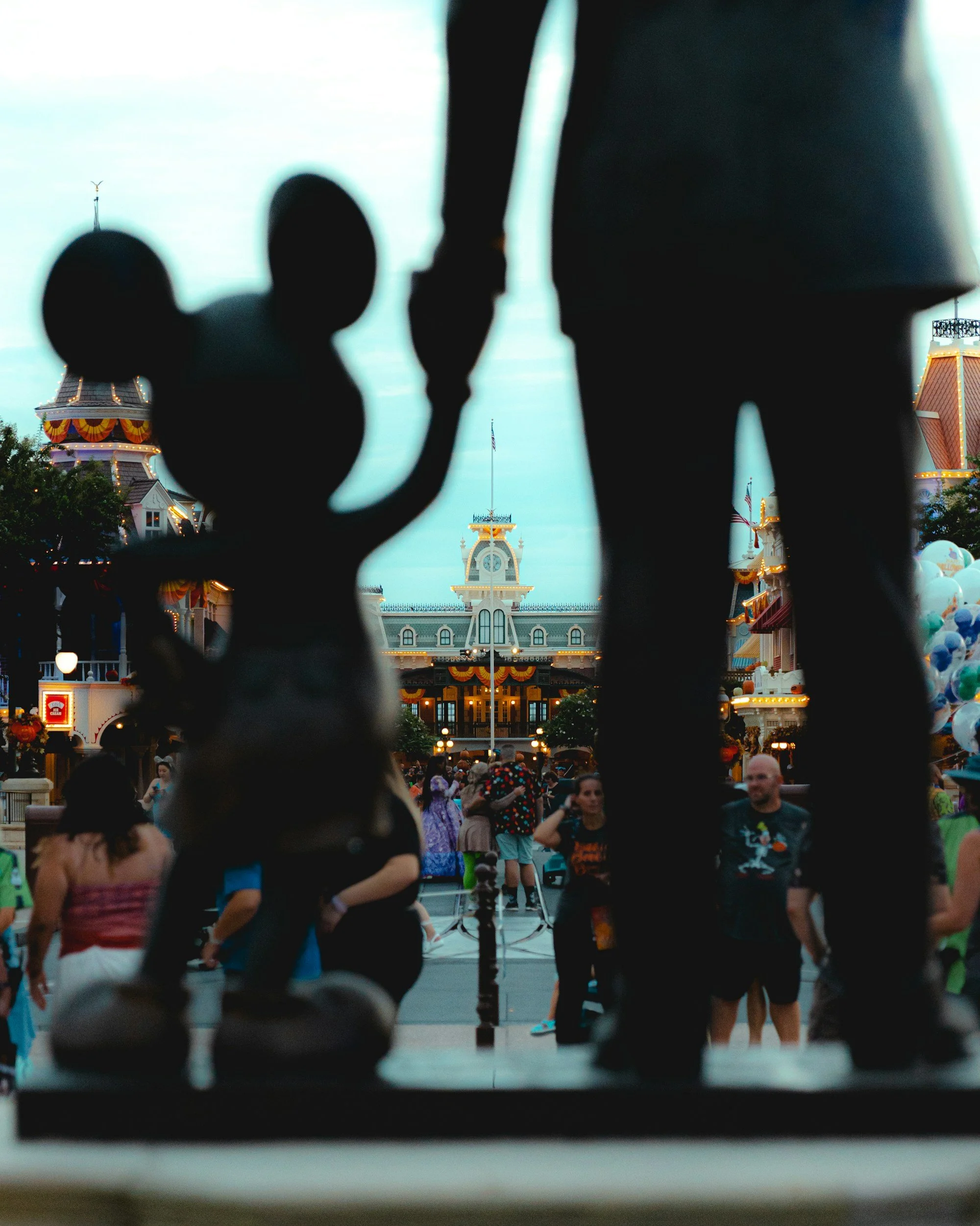 Silhouette of Mickey Mouse and a person holding hands in front of Disneyland's Main Street with the castle in the background at sunset.