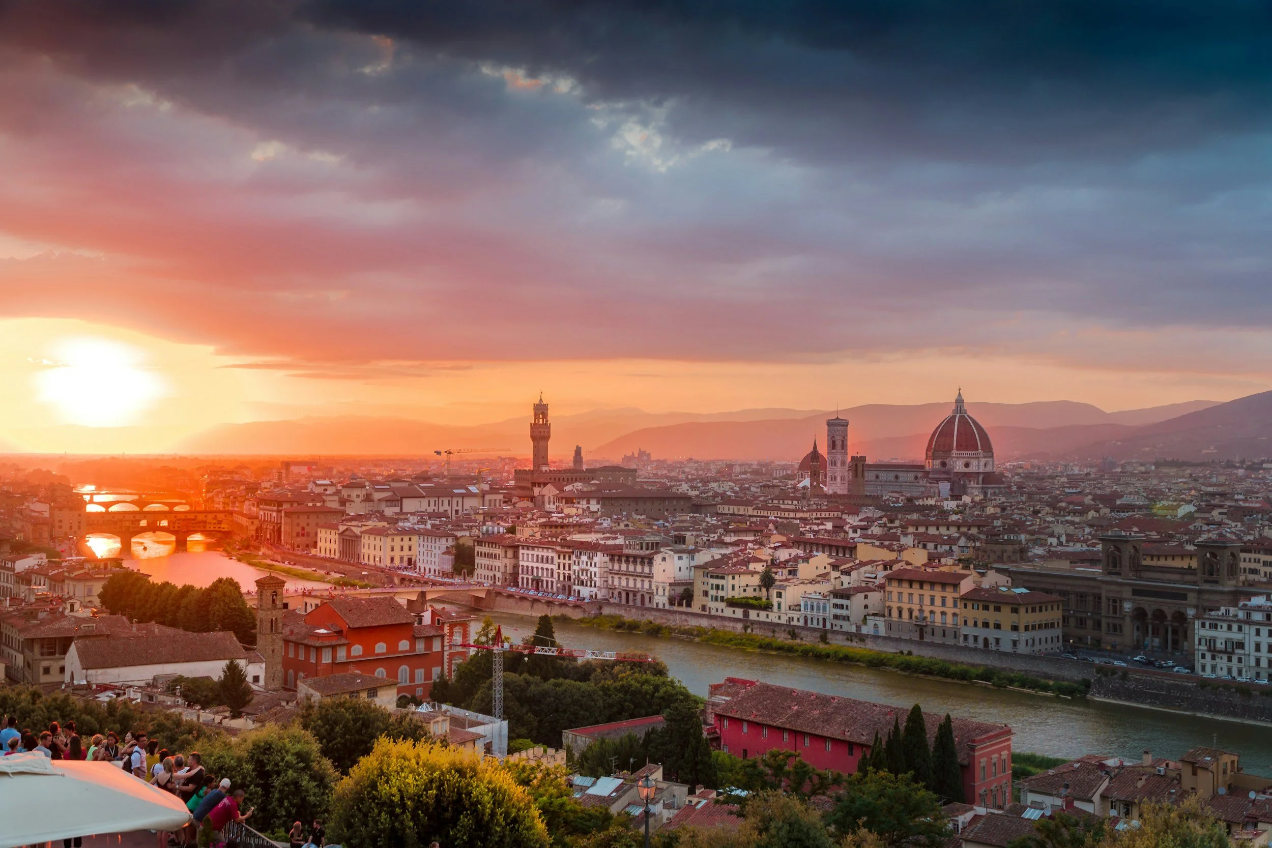 Sunset over the city of Florence, Italy, with historic buildings including the Florence Cathedral with its iconic dome, and the tower of Palazzo Vecchio along the Arno River.