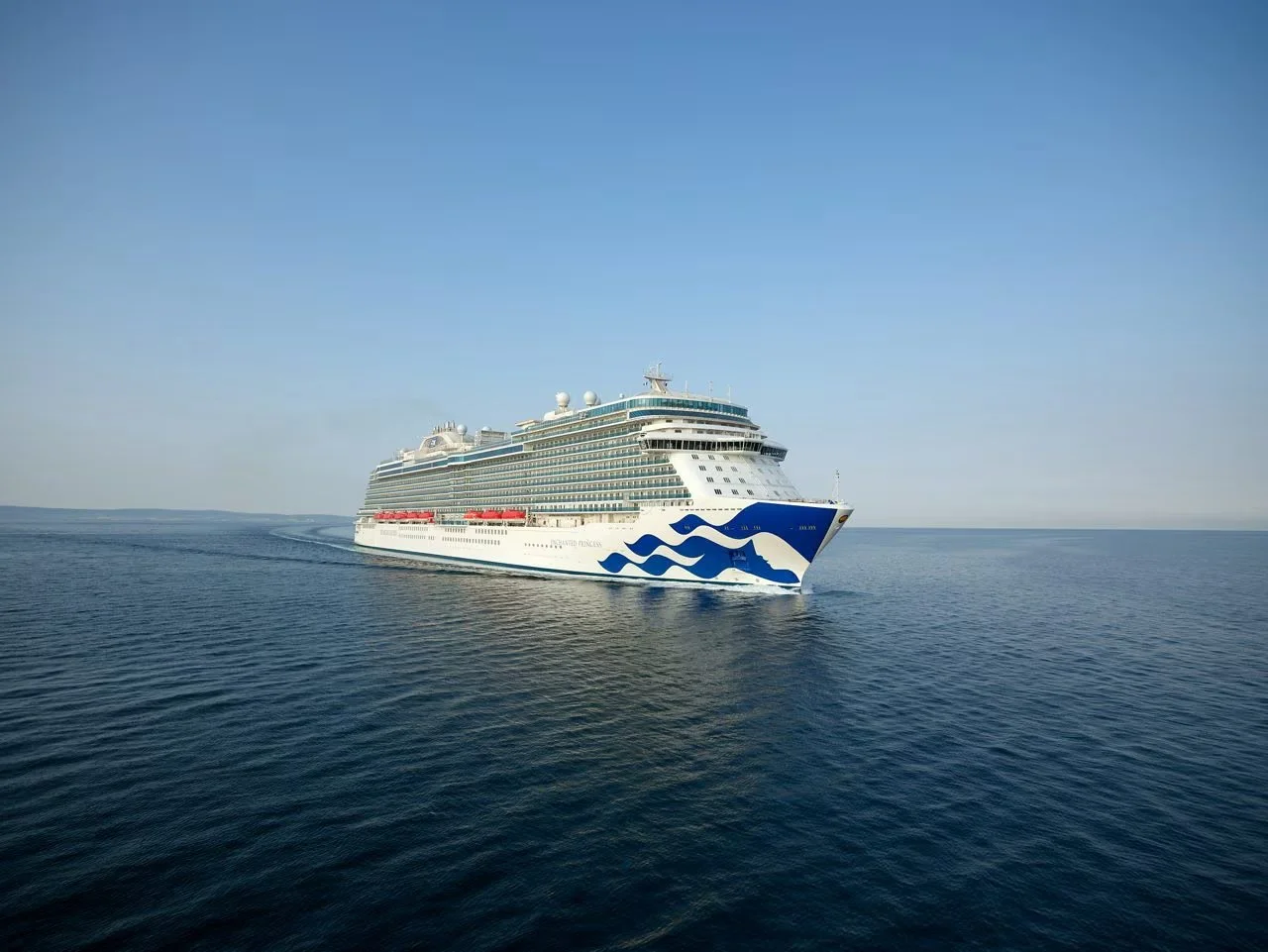 A large white cruise ship with blue wave designs on the hull sailing on calm open water under a clear blue sky.