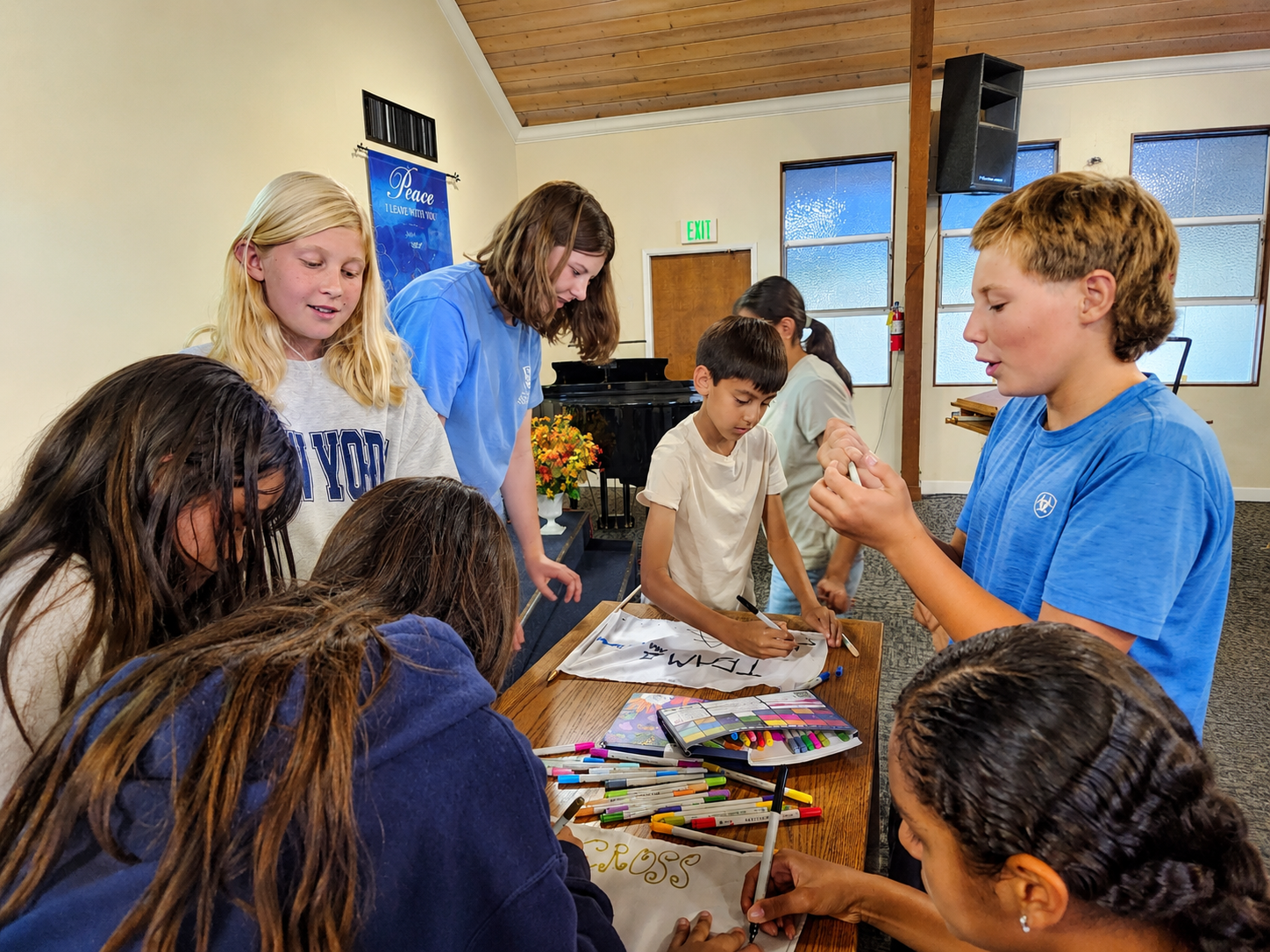 Group of children and a young woman gathered around a table, doing arts and crafts in a room with wooden ceiling, windows, and a sign that reads 'Peace I leave with you'