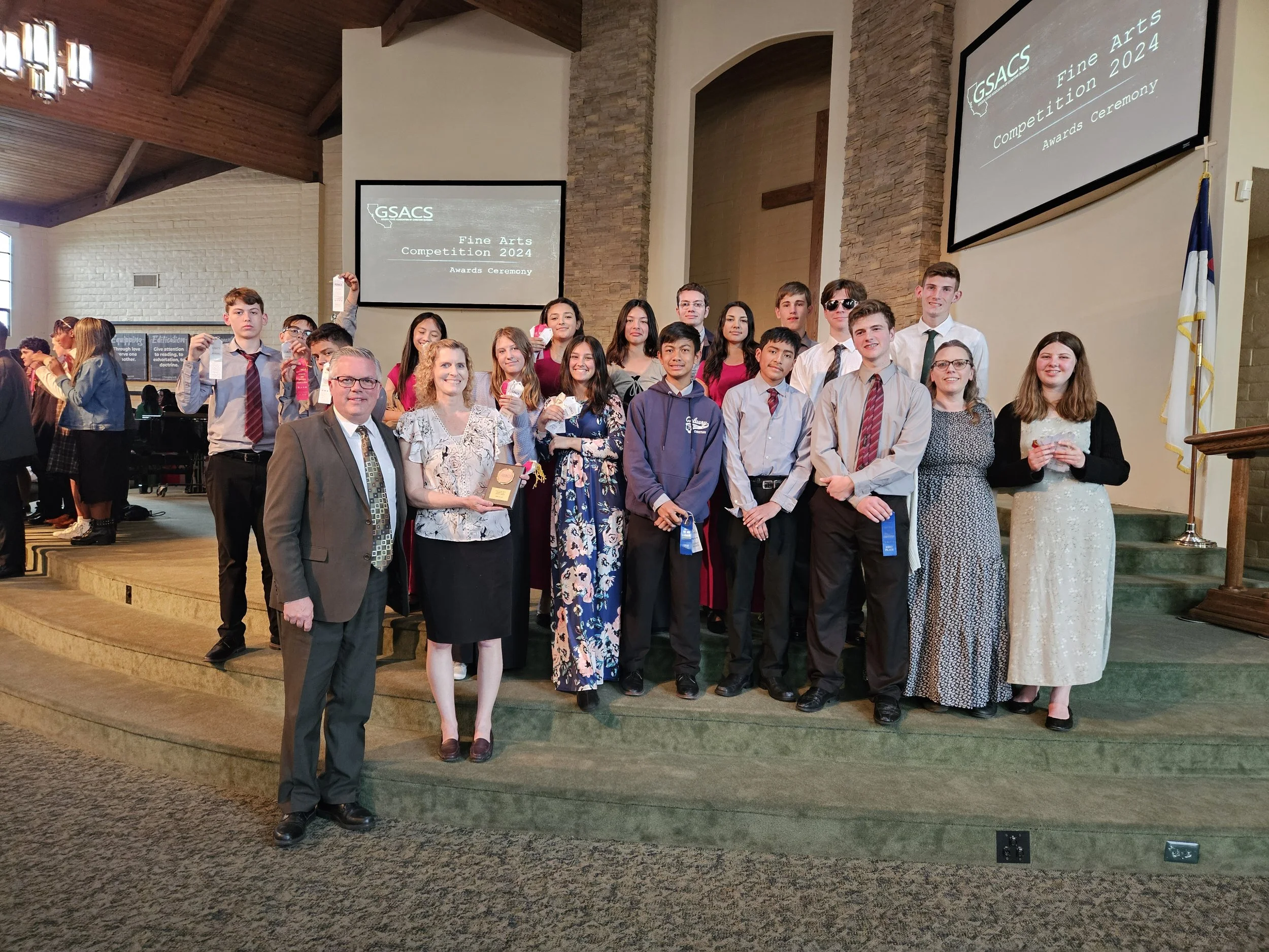Group of young students and adults standing on stage, holding awards and ribbons, at a fine arts competition awards ceremony in a church or community hall with stone walls and a high wooden ceiling.