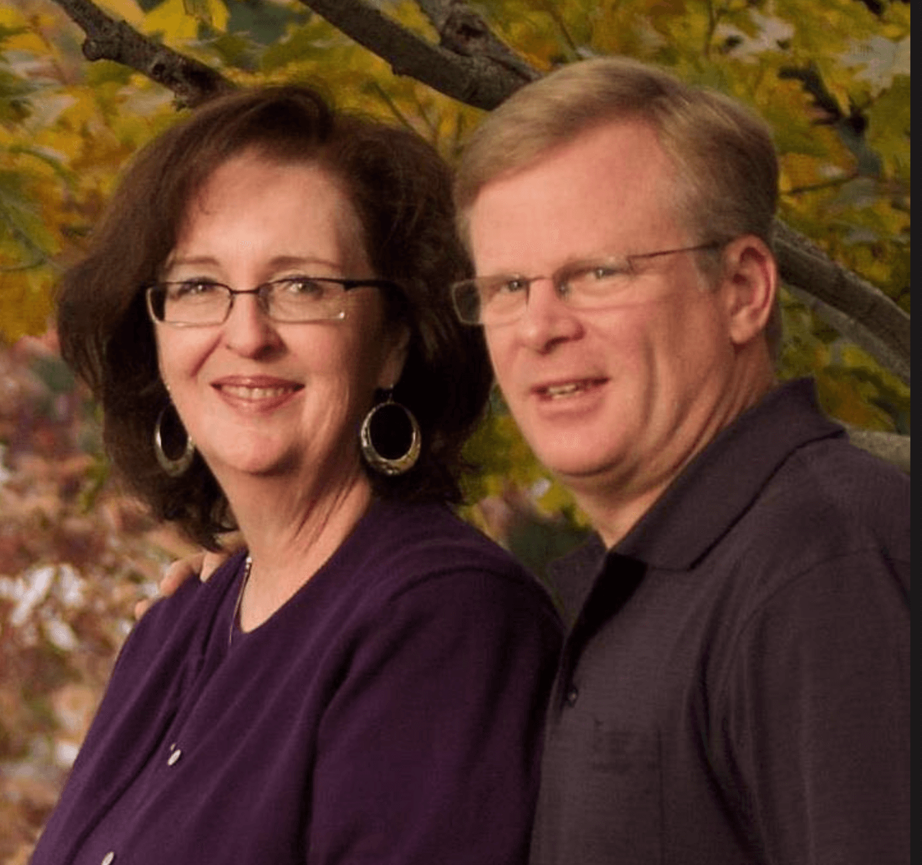 A woman with dark hair and glasses smiling, standing next to a man with light hair and glasses, both outdoors with autumn leaves in the background.