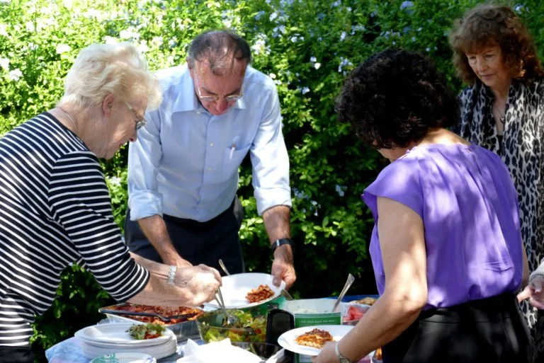Four elderly women and a man are serving and eating food outdoors at a table with various dishes, surrounded by green foliage.