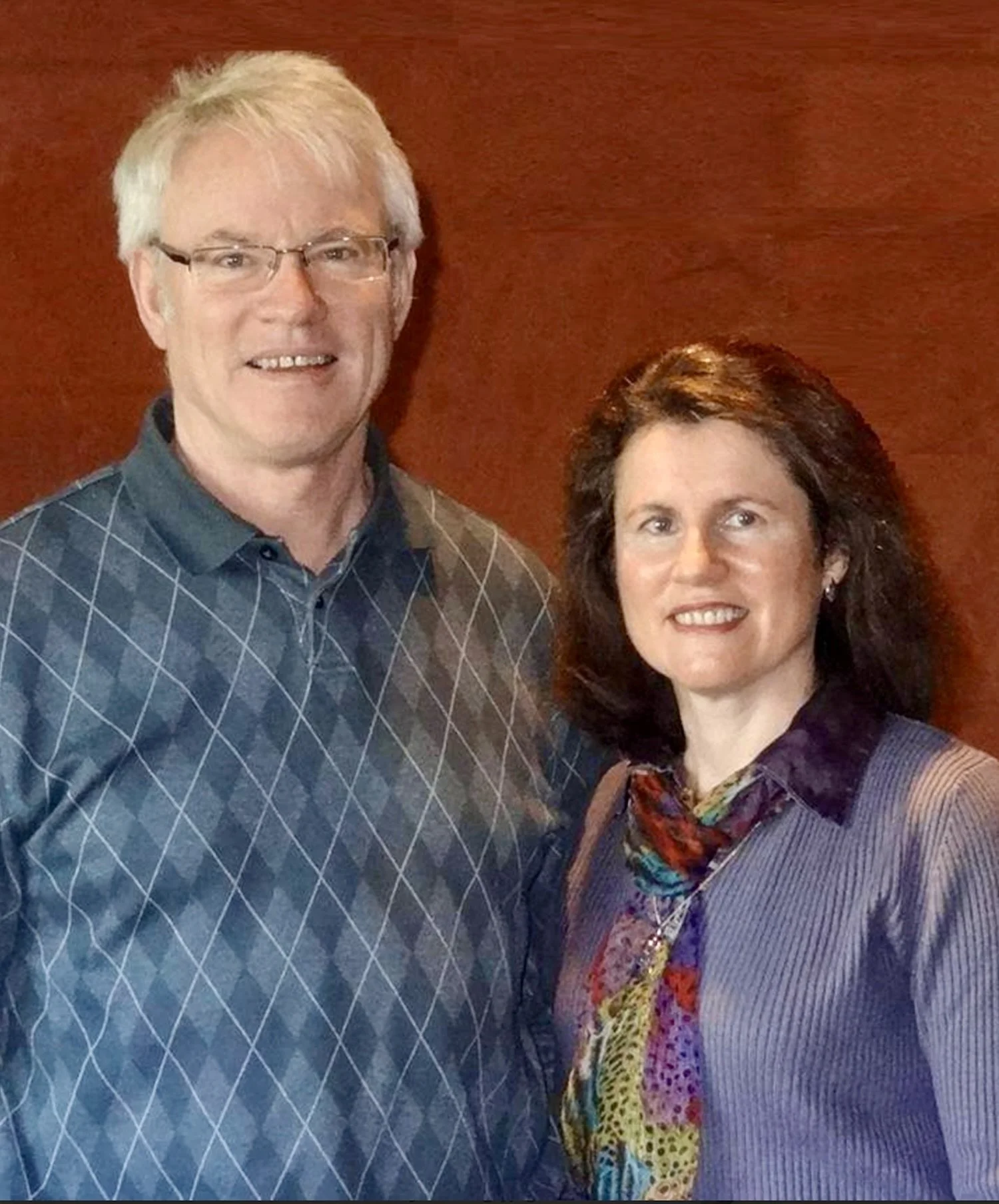 A man with gray hair and glasses and a woman with dark hair and a colorful scarf standing in front of a wooden wall.